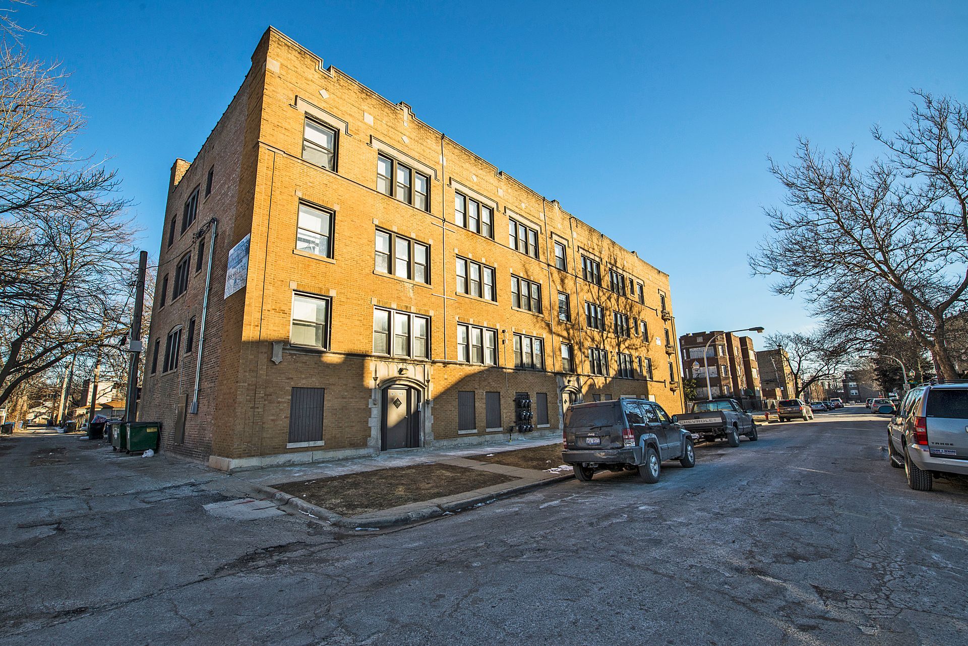 Four-story brick apartment building on a city street. Cars parked on the side. Clear, sunny sky.