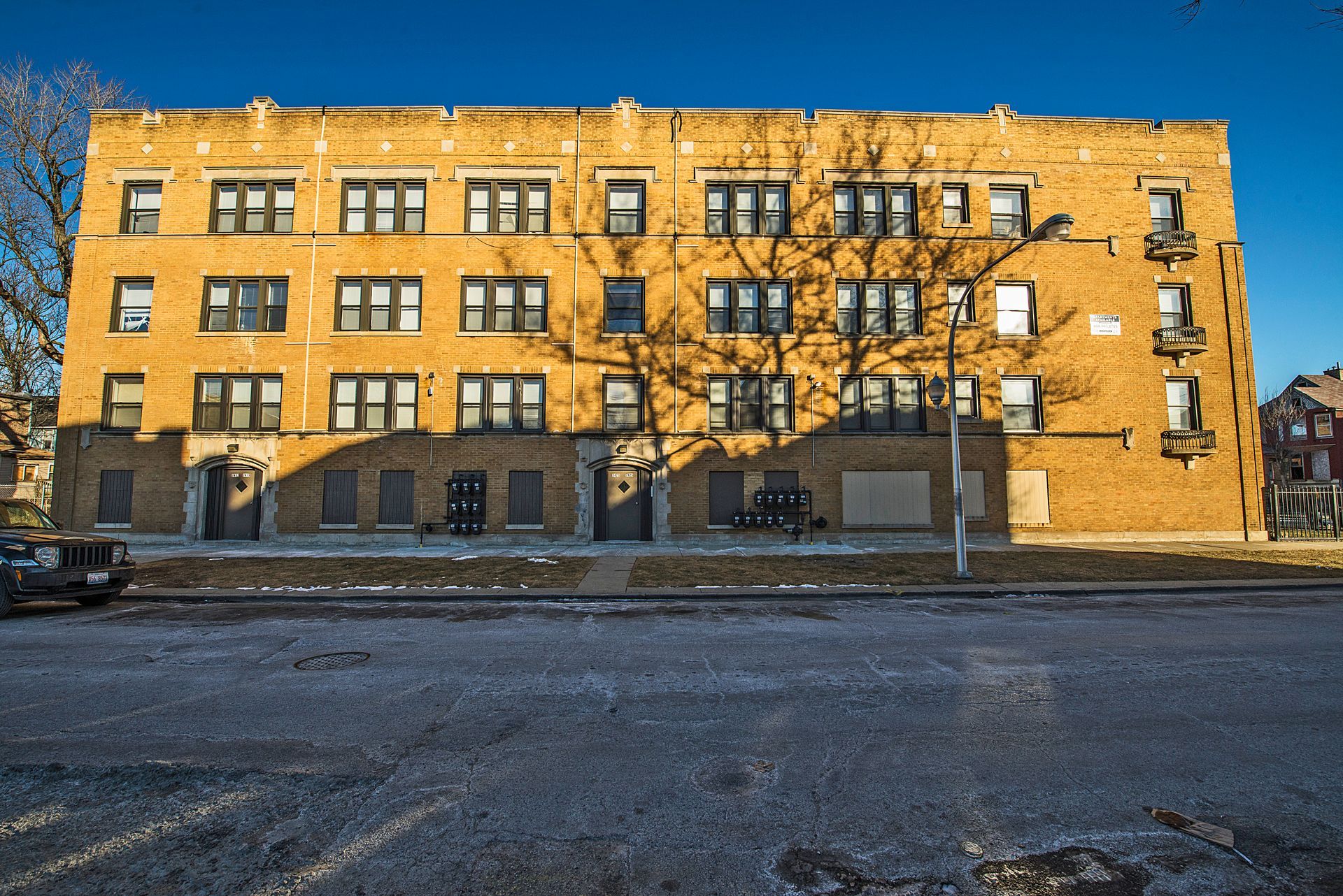 Three-story brick apartment building on a winter day with bare trees casting shadows.
