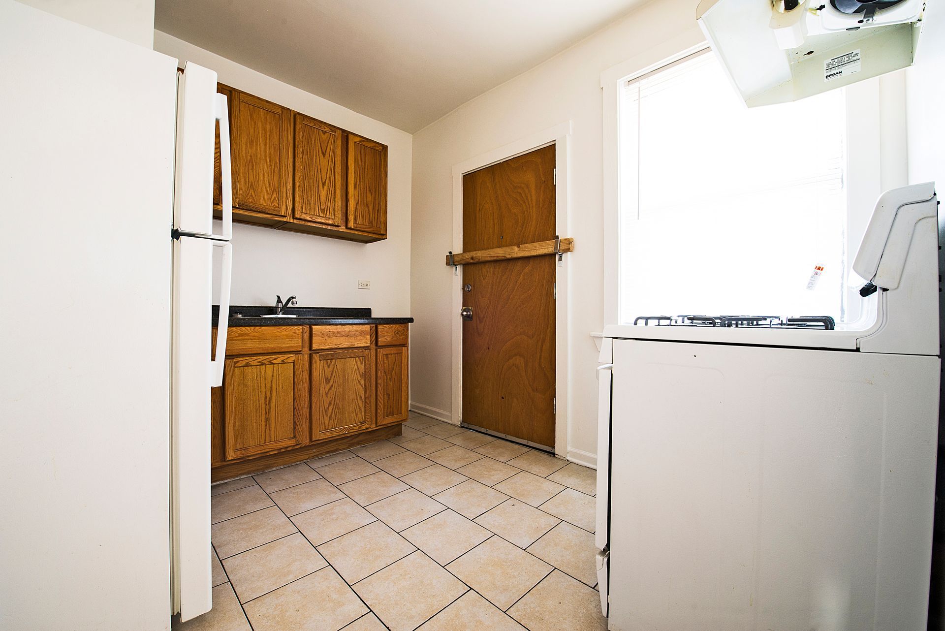 Empty kitchen with white appliances, wooden cabinets, and beige tiled floor.