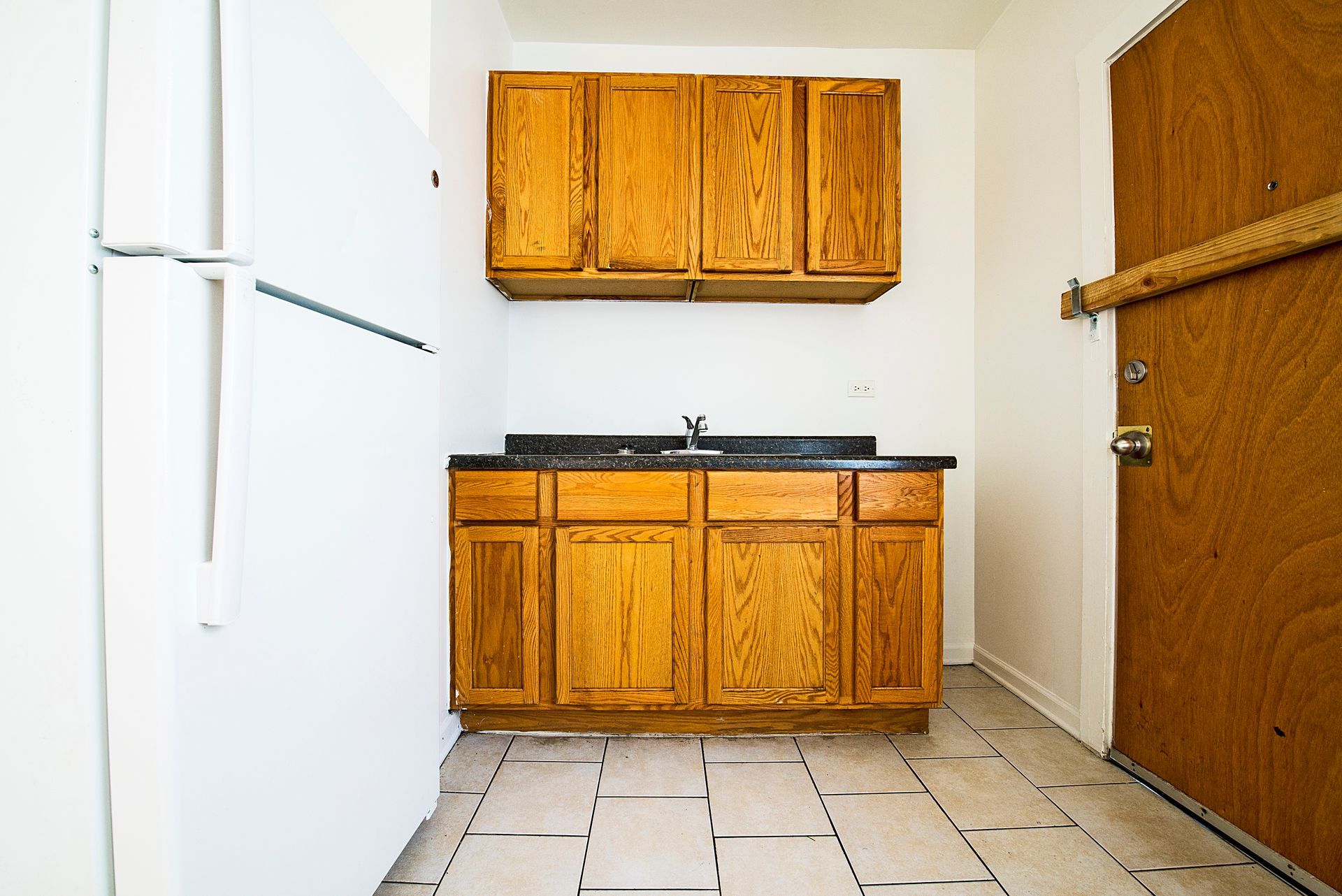 Small kitchen with wood cabinets, countertop, sink, and fridge. Doorway on the right. Tiled floor.