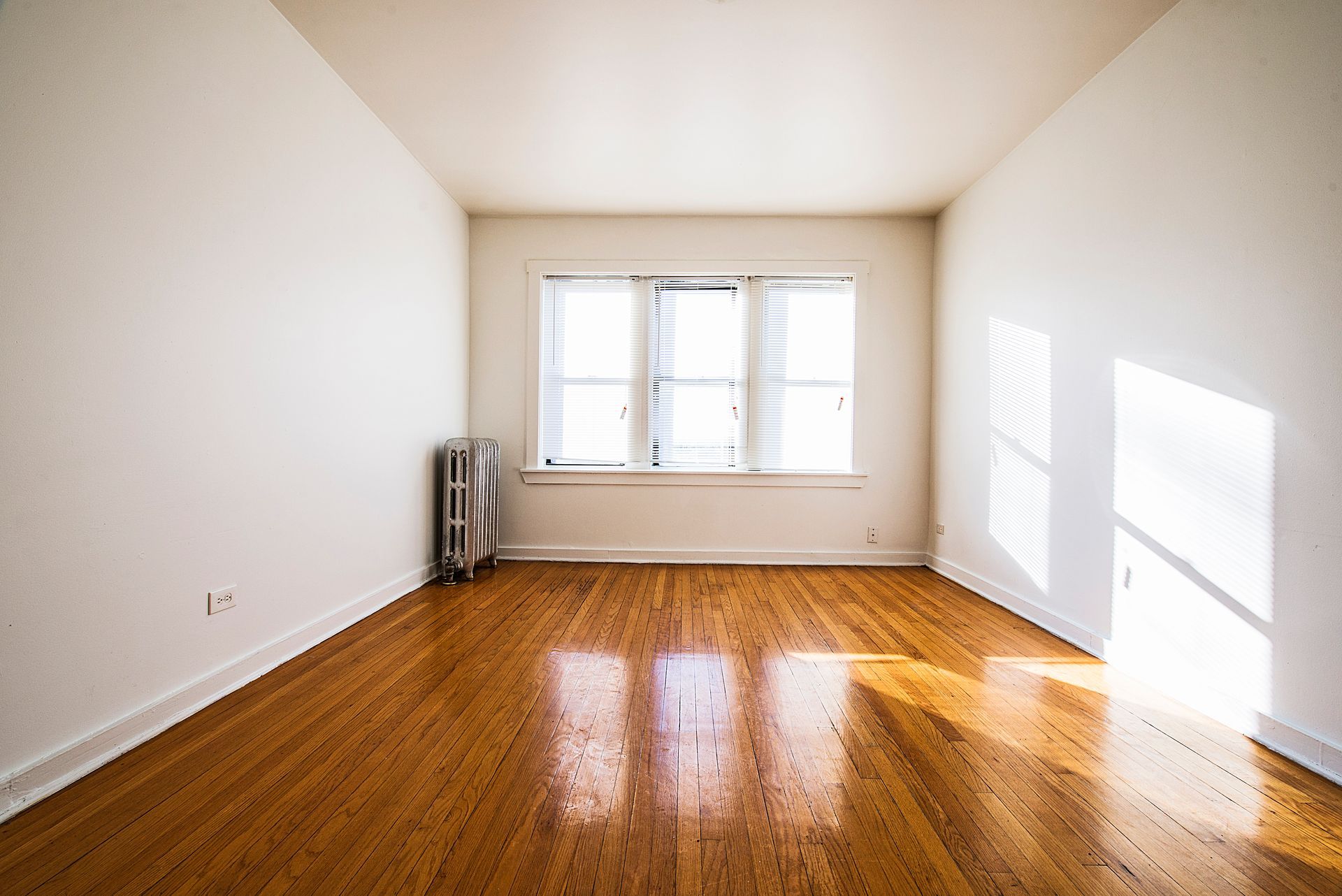 Empty room with wooden floor, window, and radiator. White walls. Sunlight streams in.