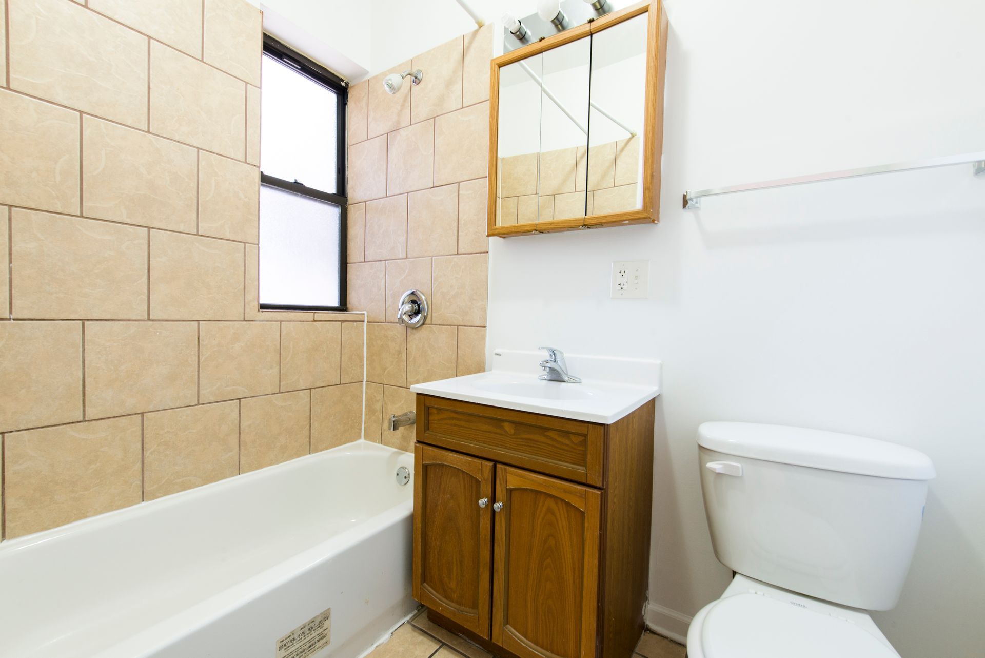 Bathroom with a bathtub, toilet, vanity, and window. Beige tile, wood cabinet, and white fixtures.