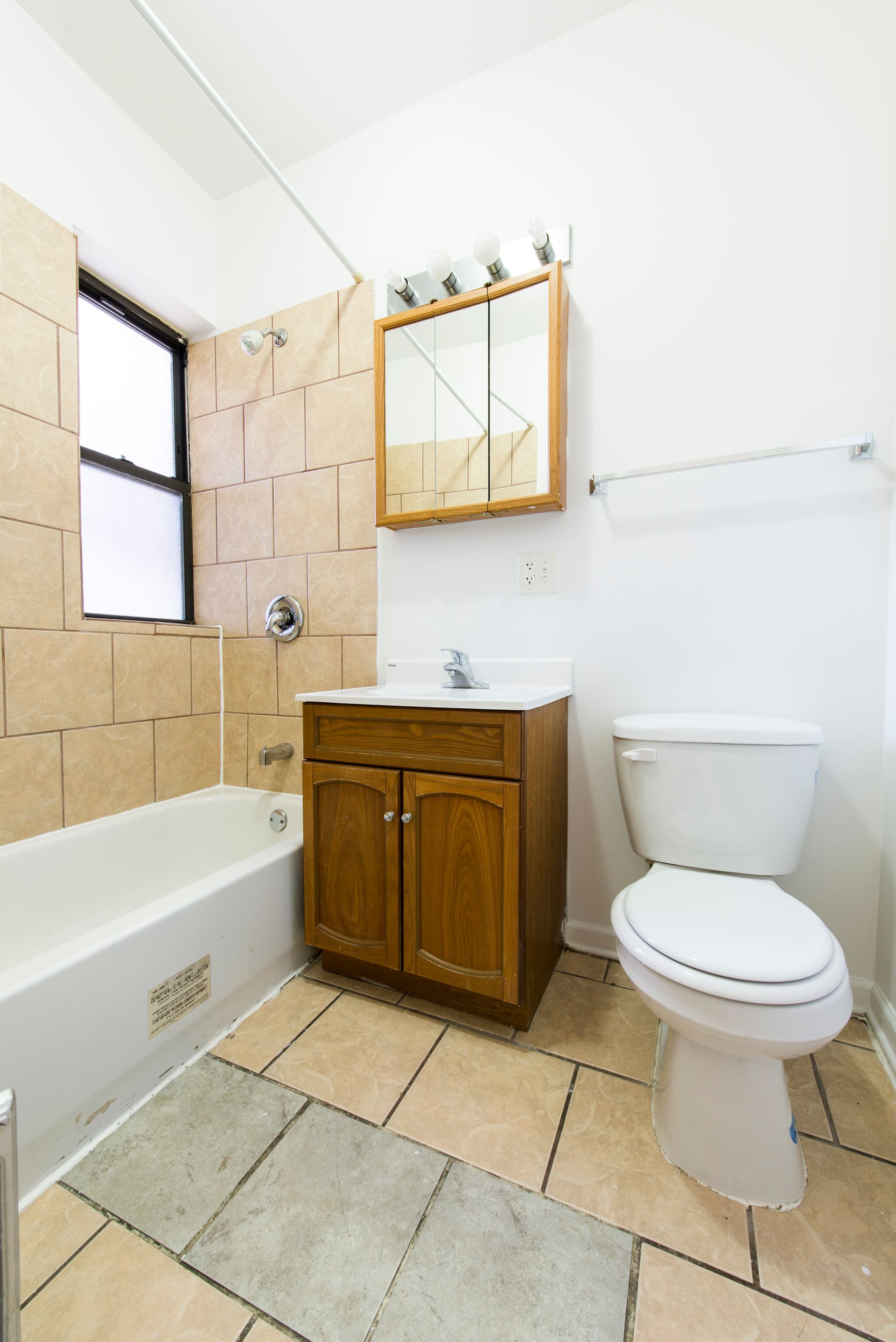 Bathroom with bathtub, toilet, vanity, and mirror. Beige tiles on walls and floor. Window on the left.
