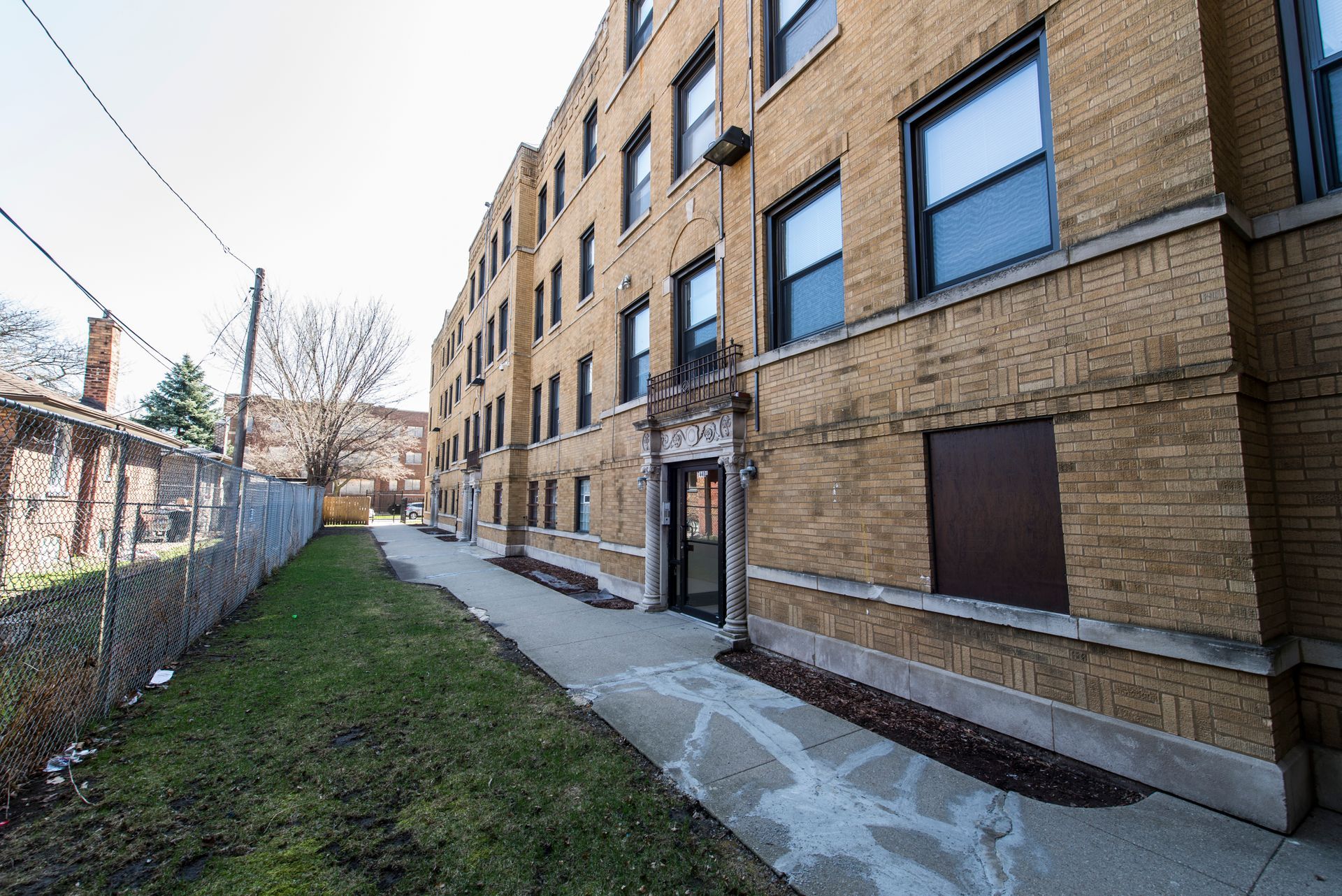 A brick apartment building with a narrow walkway, grass, and a chain-link fence.