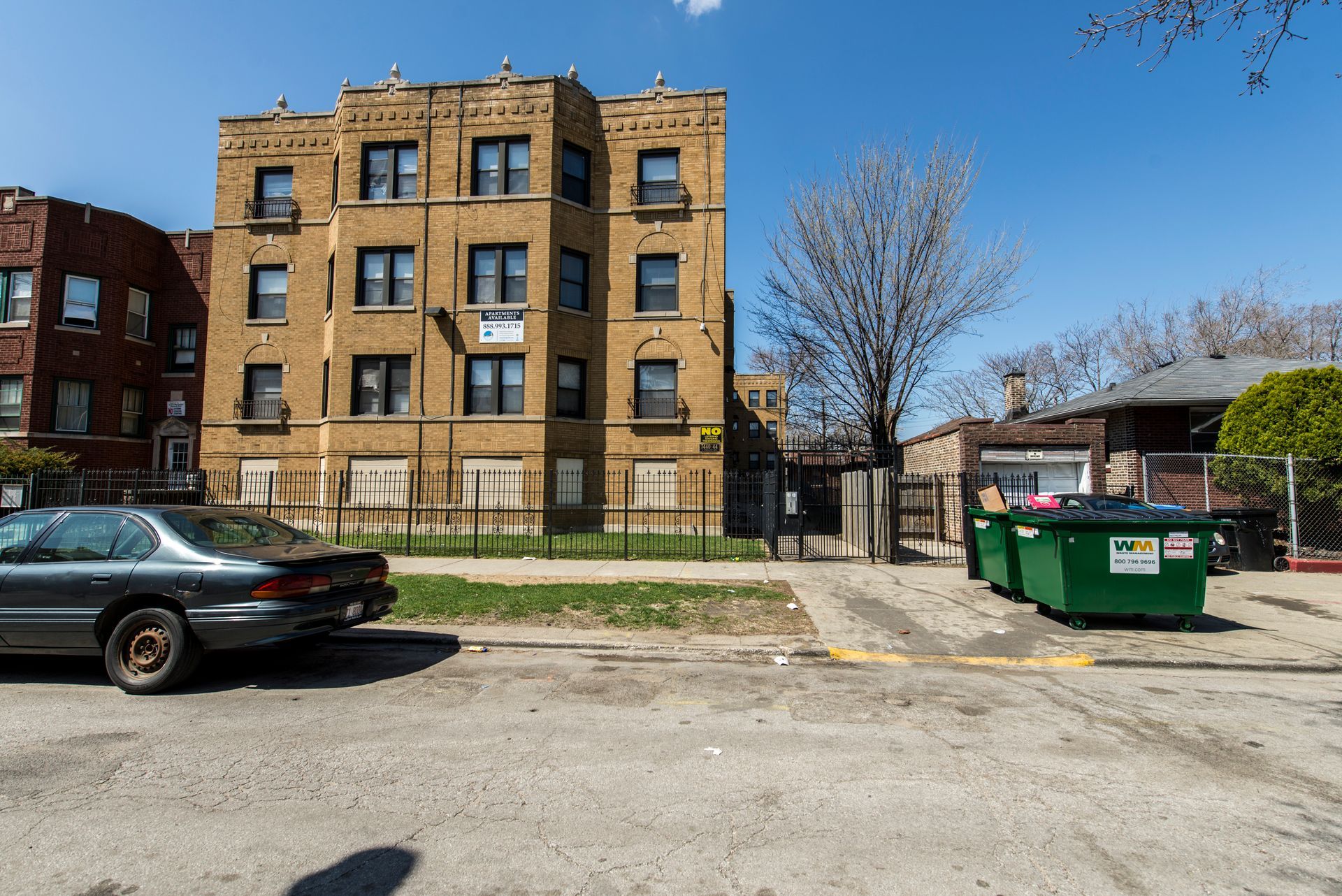 A tan apartment building on a city street, boarded-up windows, a car parked nearby, green dumpsters.