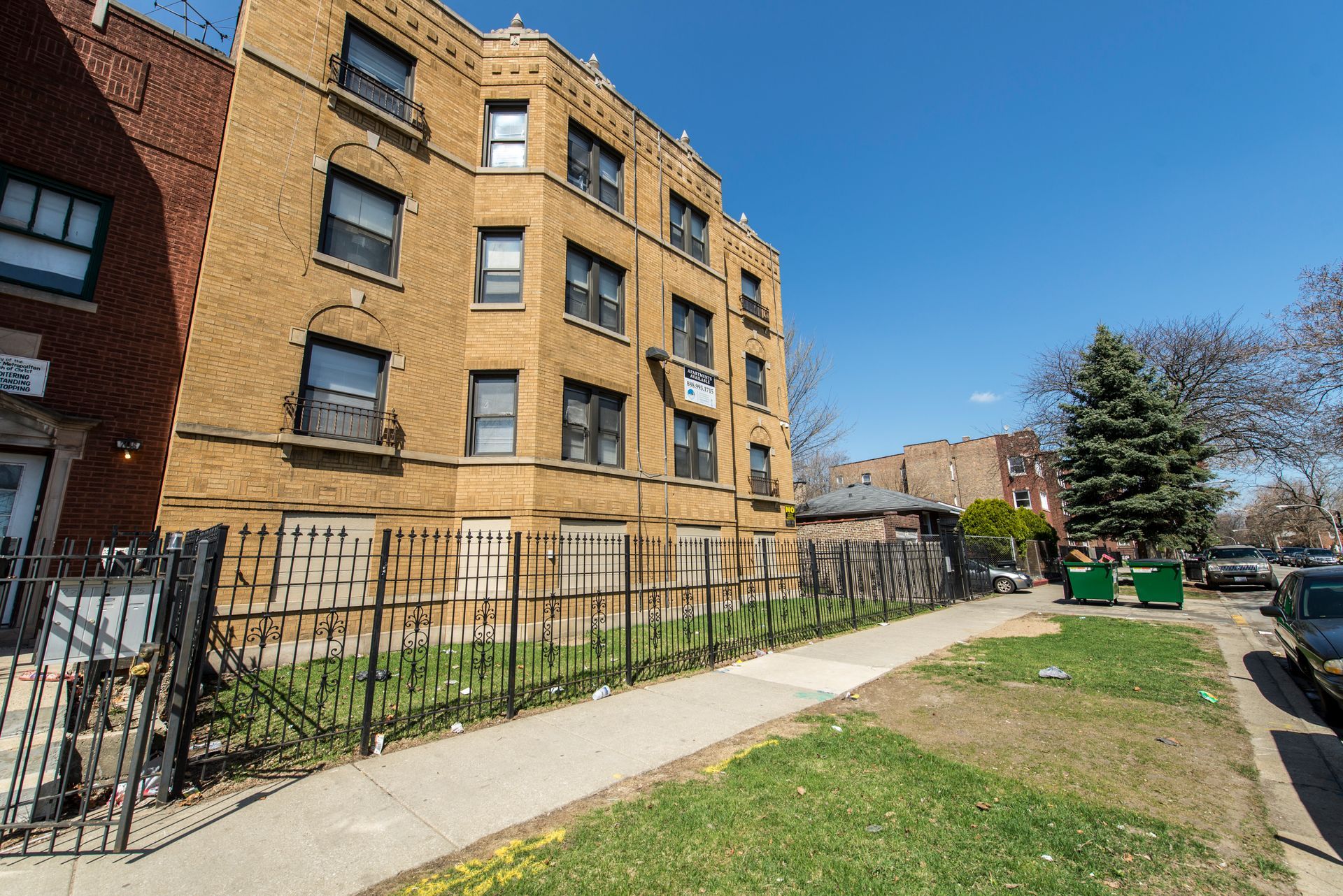 Yellow brick apartment building with black fence and sidewalk on a sunny day.