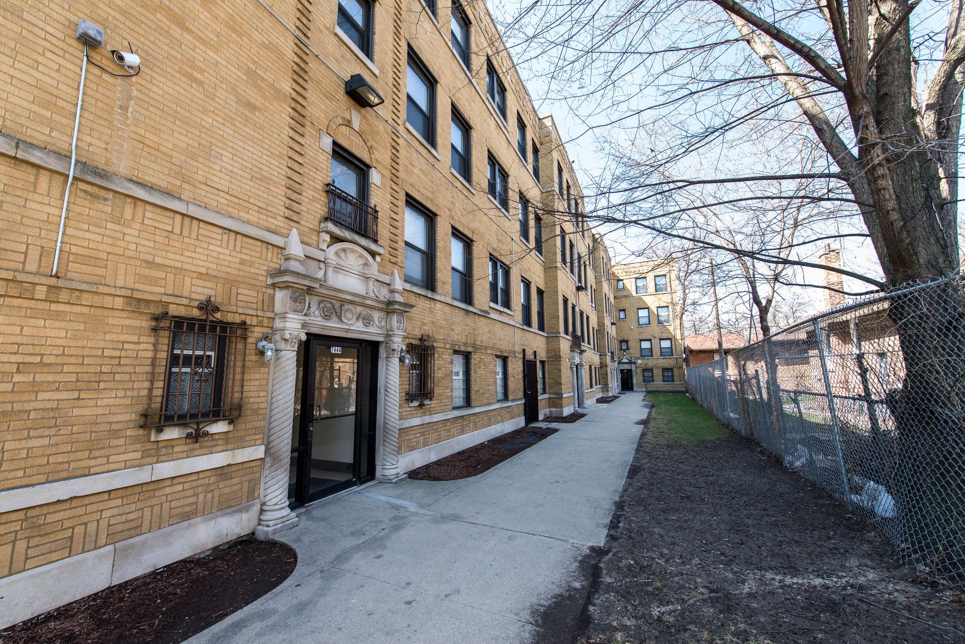 Yellow brick apartment building with a narrow walkway, trees, and chain-link fence.