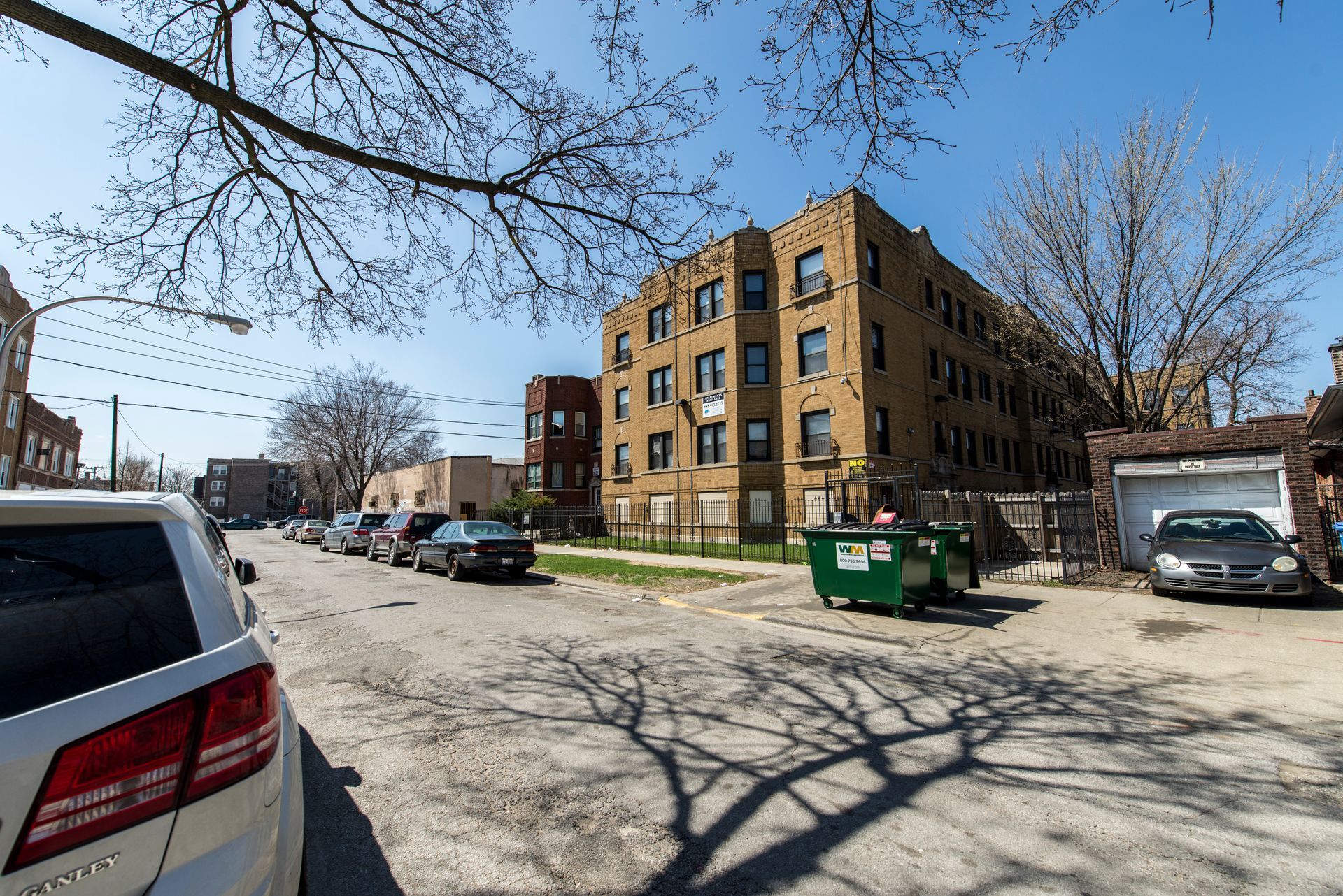Apartment building on a sunny street, cars parked, green dumpster, bare tree branches overhead.