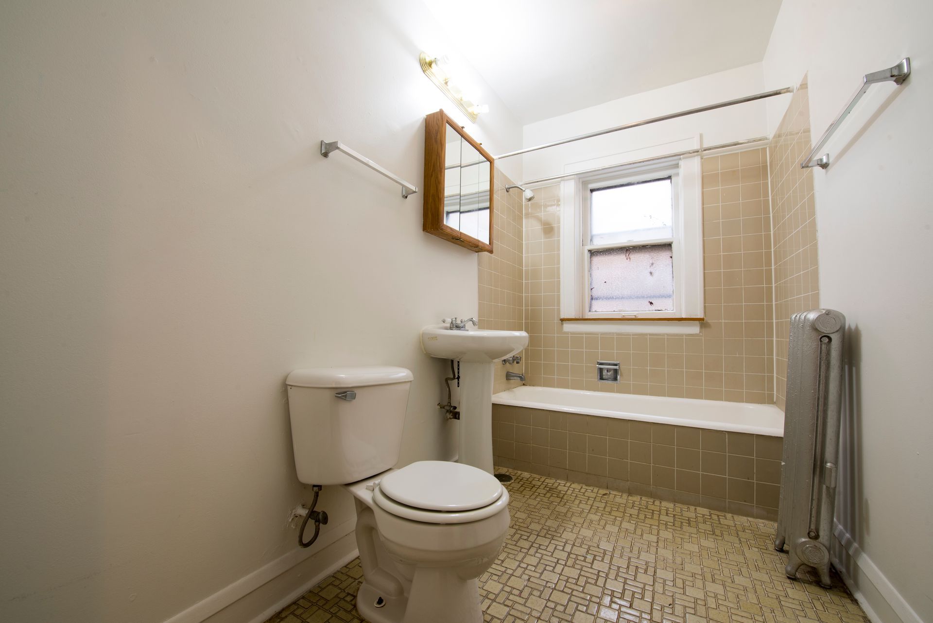 Bathroom with toilet, sink, tub, window, and radiator. Beige tile and walls, light fixtures.