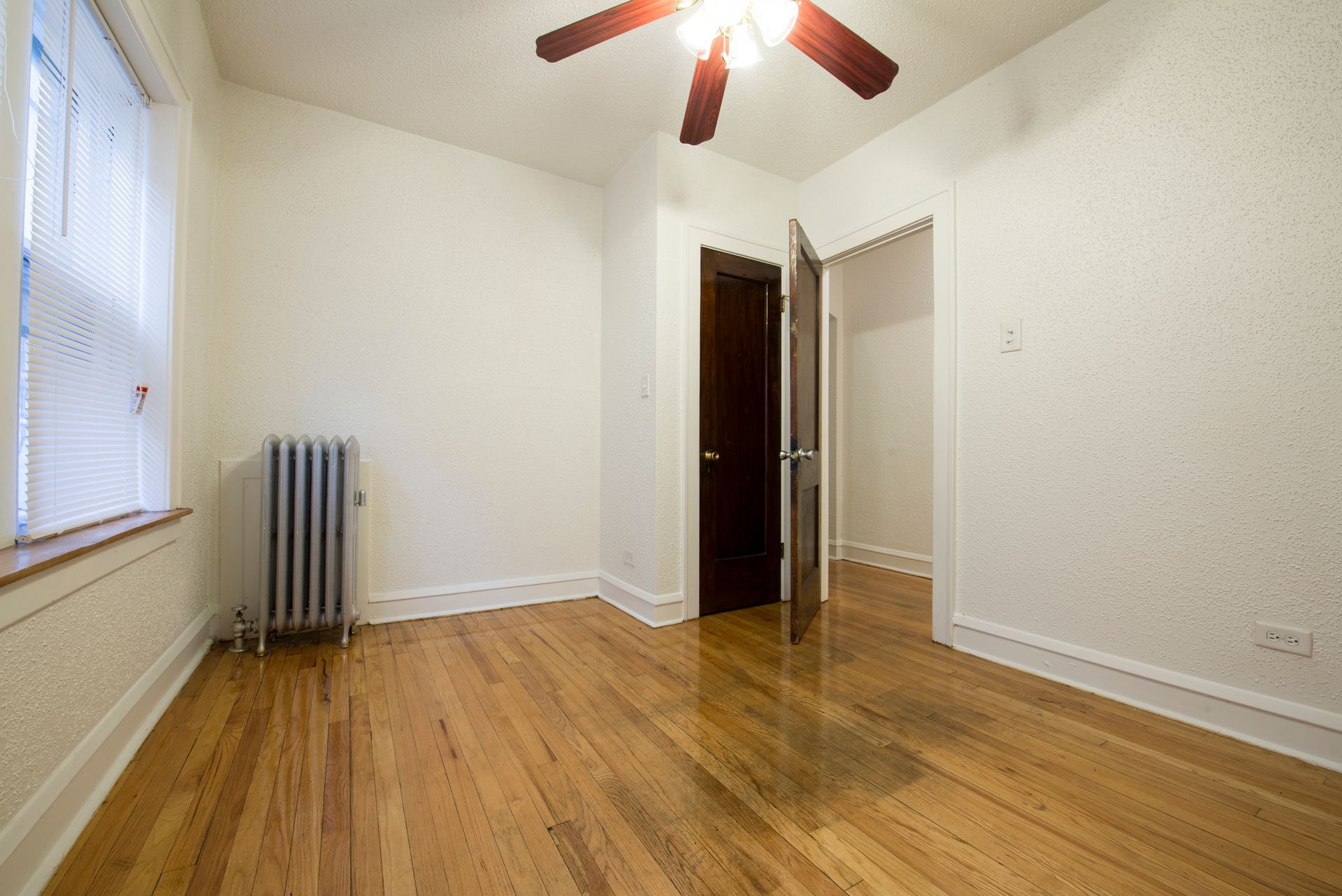 Empty room with hardwood floors, radiator, window, dark door, ceiling fan.