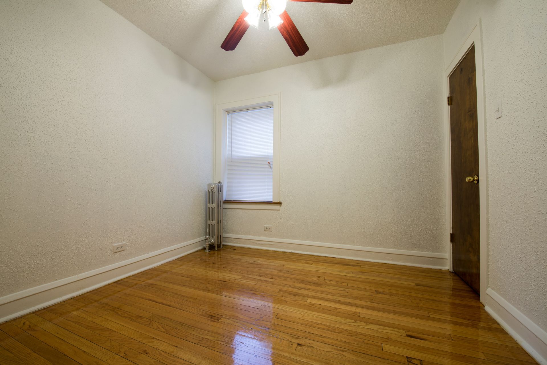 Empty room with wood floor, white walls, small window, dark brown door, ceiling fan.