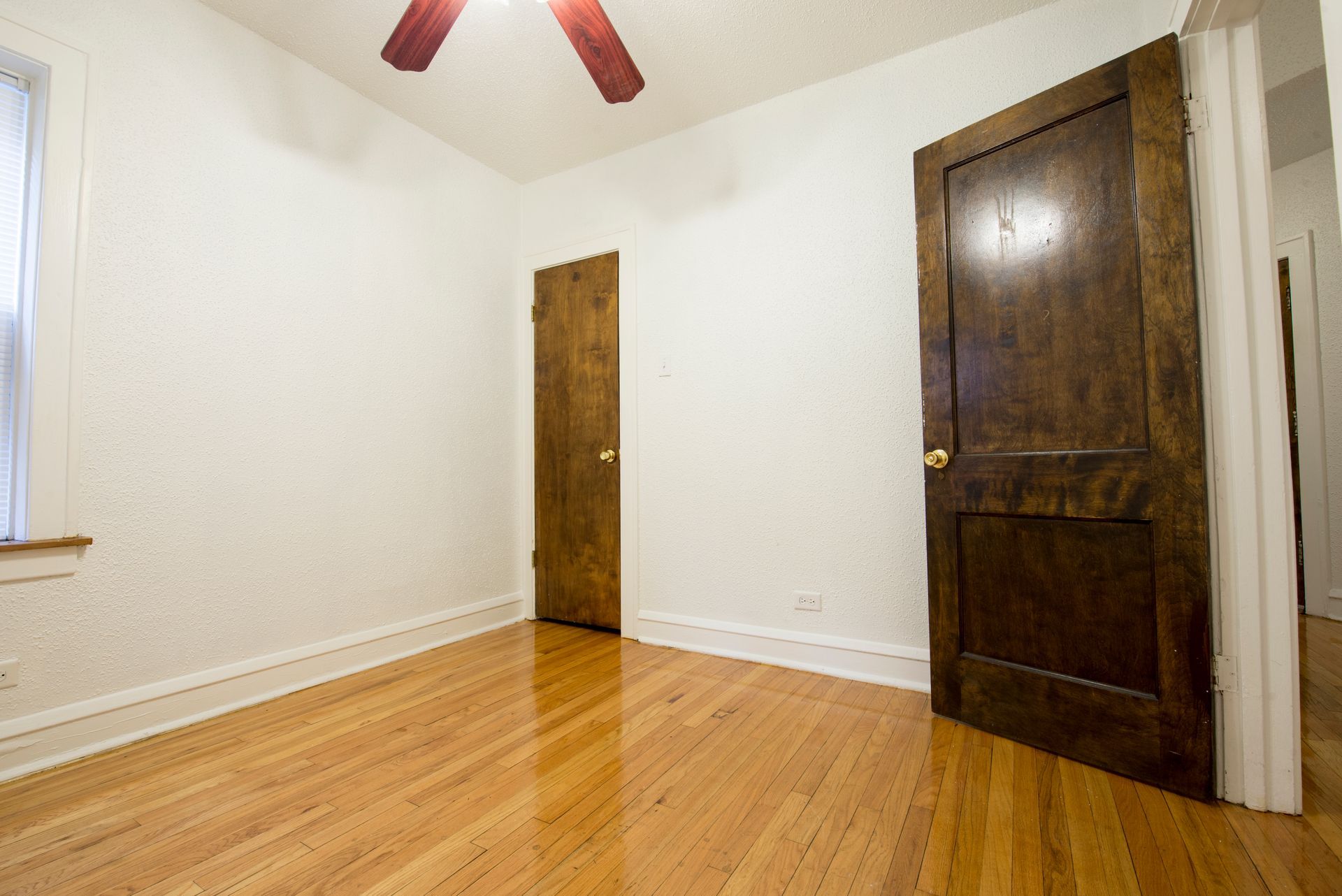Empty room with hardwood floors, white walls, two brown doors, and a ceiling fan.