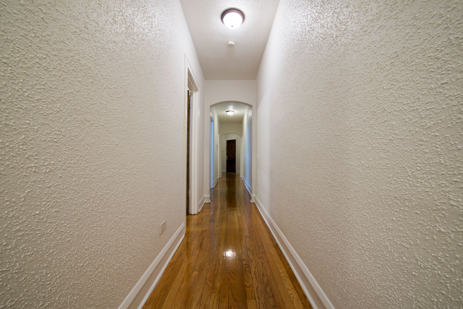 Narrow hallway with textured white walls, wooden floor, and arched doorway.