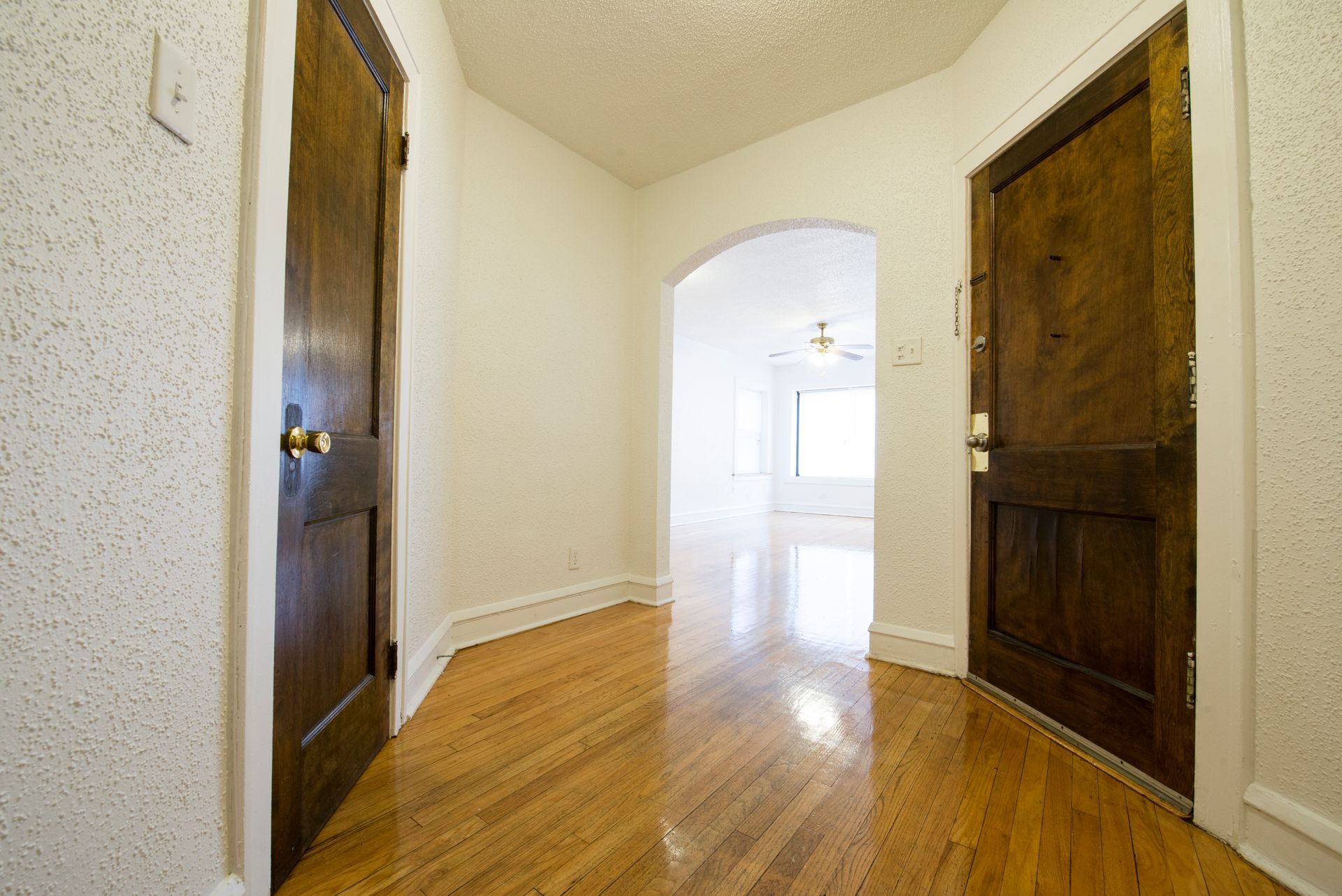 Hallway with hardwood floor, two brown doors, archway to a bright room.