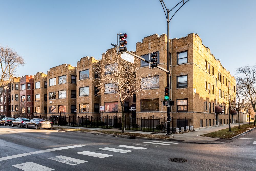 Multi-story brick apartment building on a city street corner; cars parked along the curb; traffic light overhead.