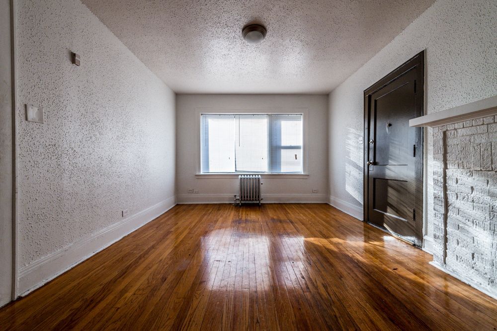 Empty room with wood floor, white textured walls, window with blinds, and a fireplace.