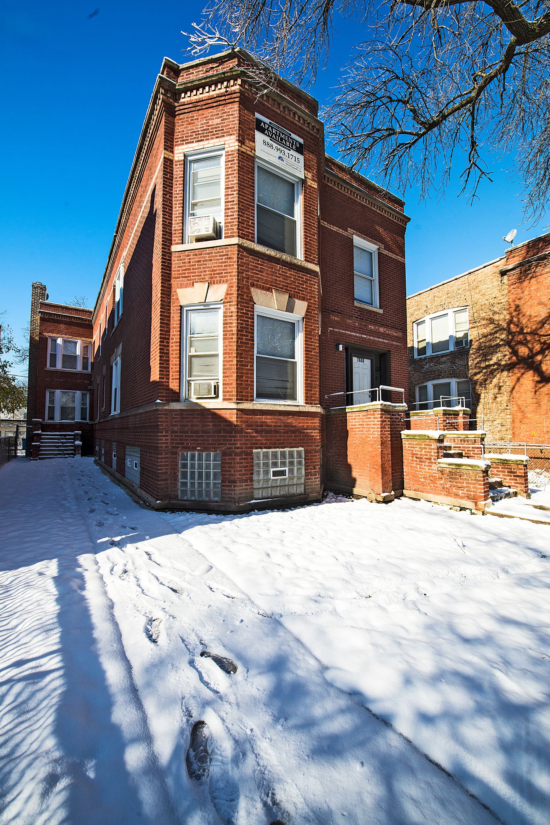 Red brick two-story building in a snowy setting with foot tracks. Blue sky in the background.