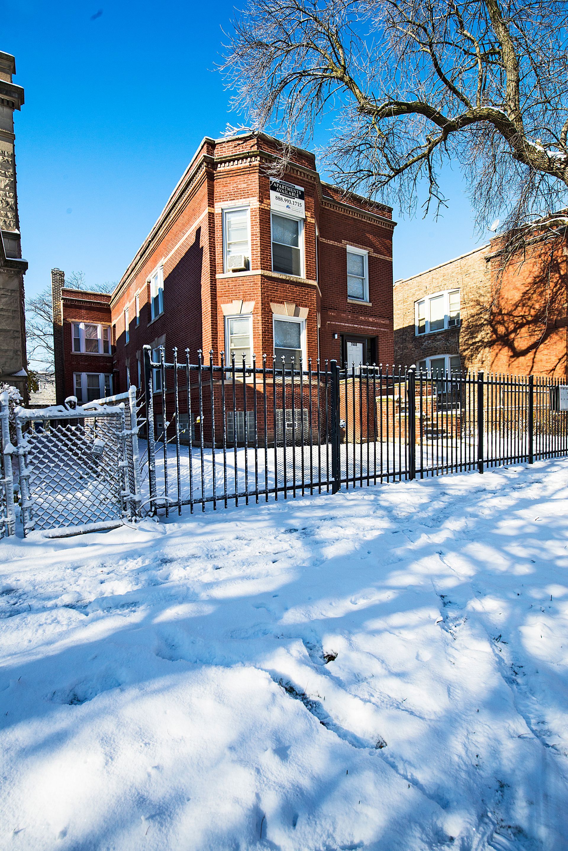 Brick apartment building with snow-covered yard and black metal fence under a clear blue sky.