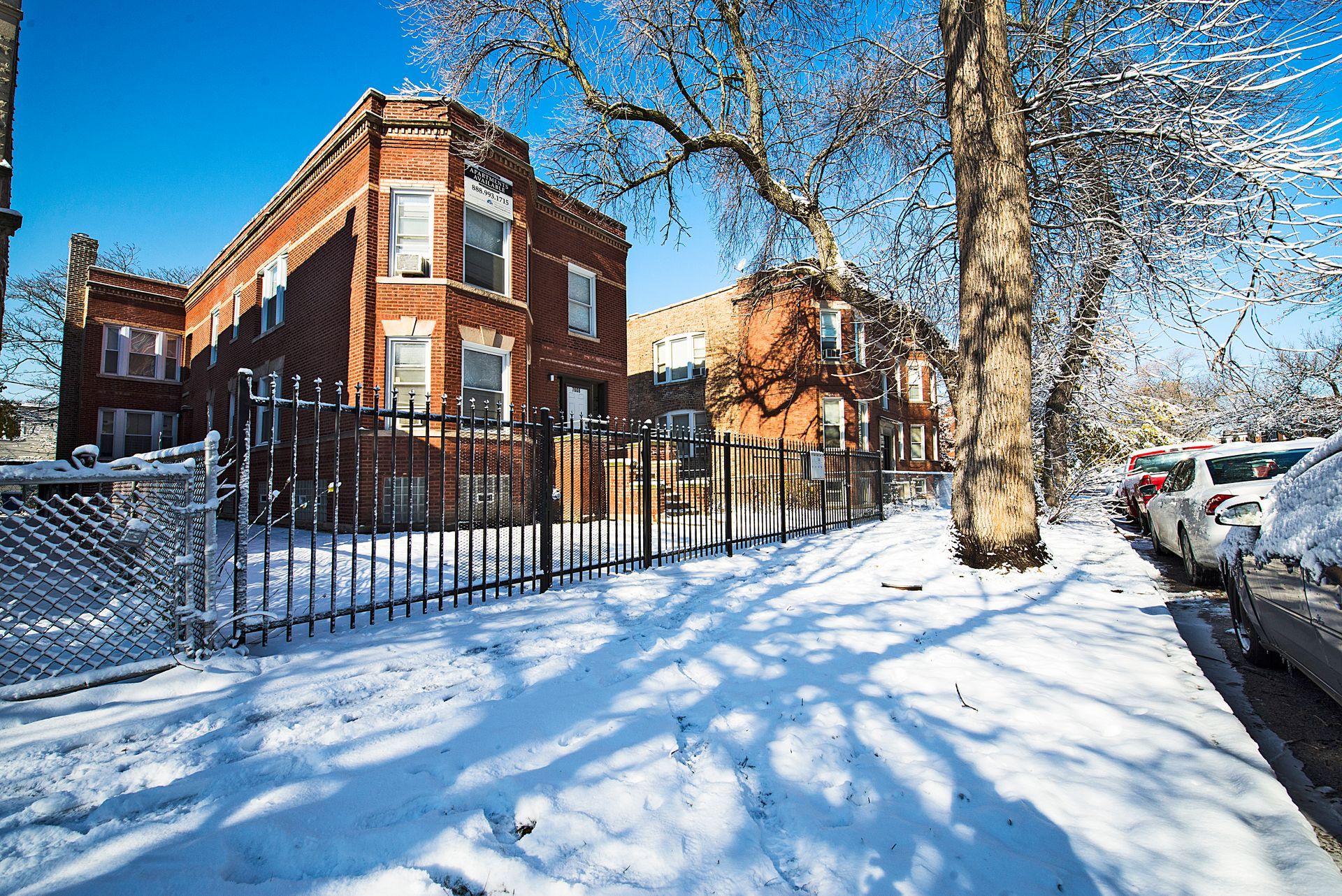 Brick apartment building in winter, surrounded by snow and a fence. Street with parked cars.