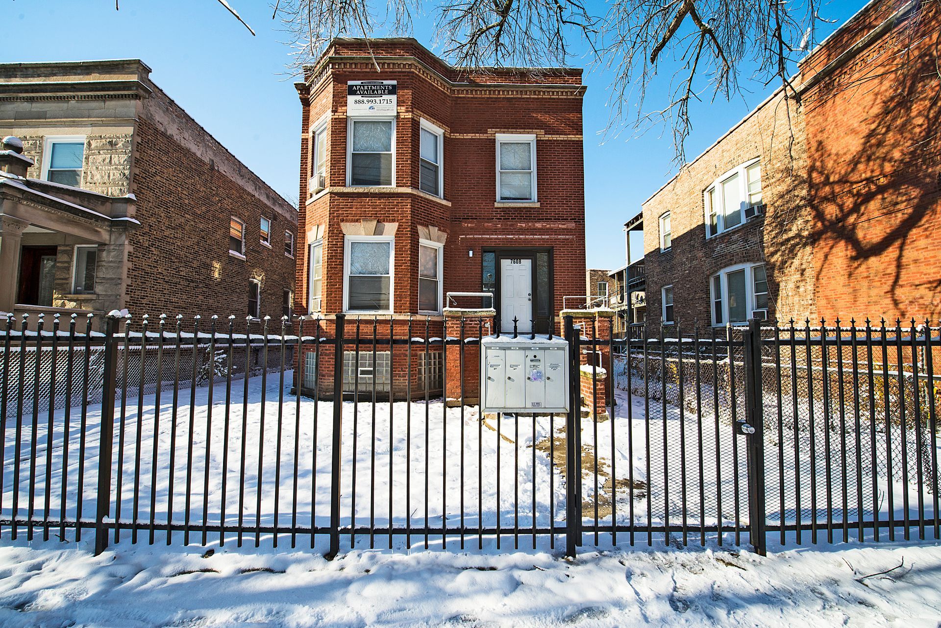 Red brick building with a black fence in front, snow on the ground, and two adjacent buildings.