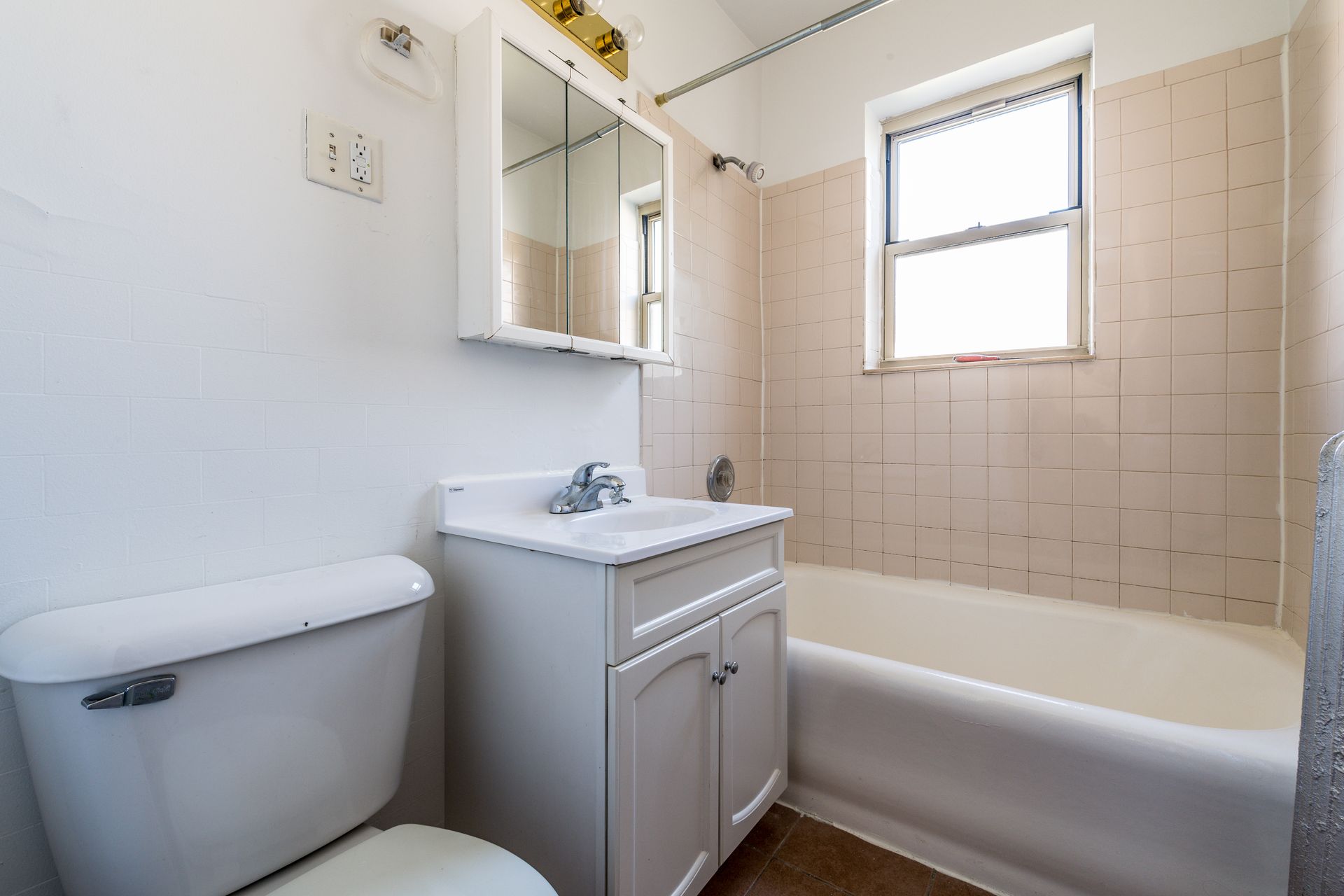 Small white bathroom with toilet, vanity, and bathtub. Beige tiled walls, small window.
