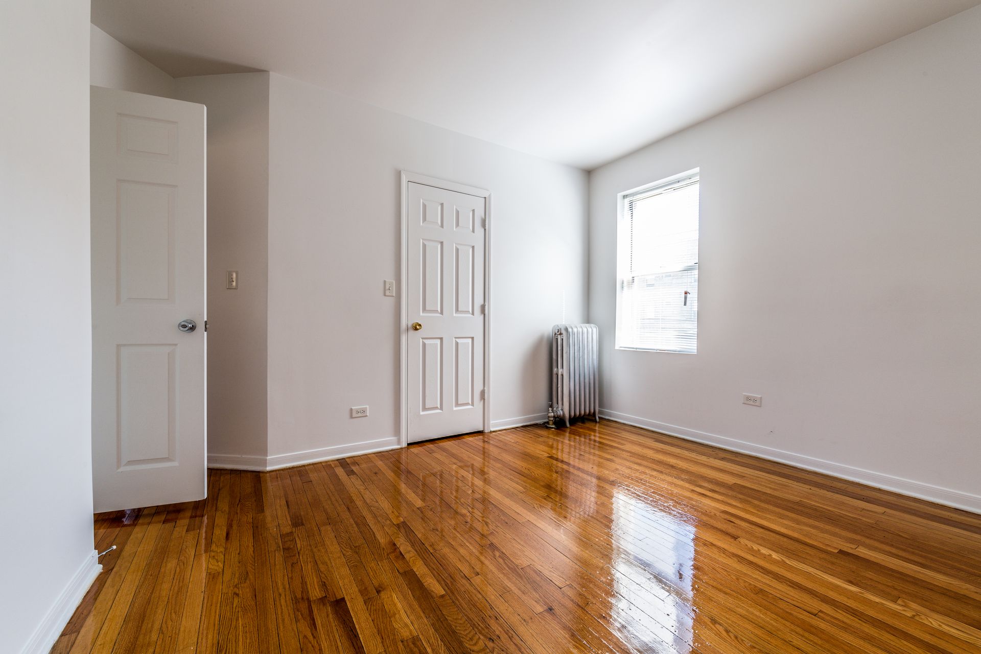 Empty room with wood floors, white walls, two closed doors, and a window.
