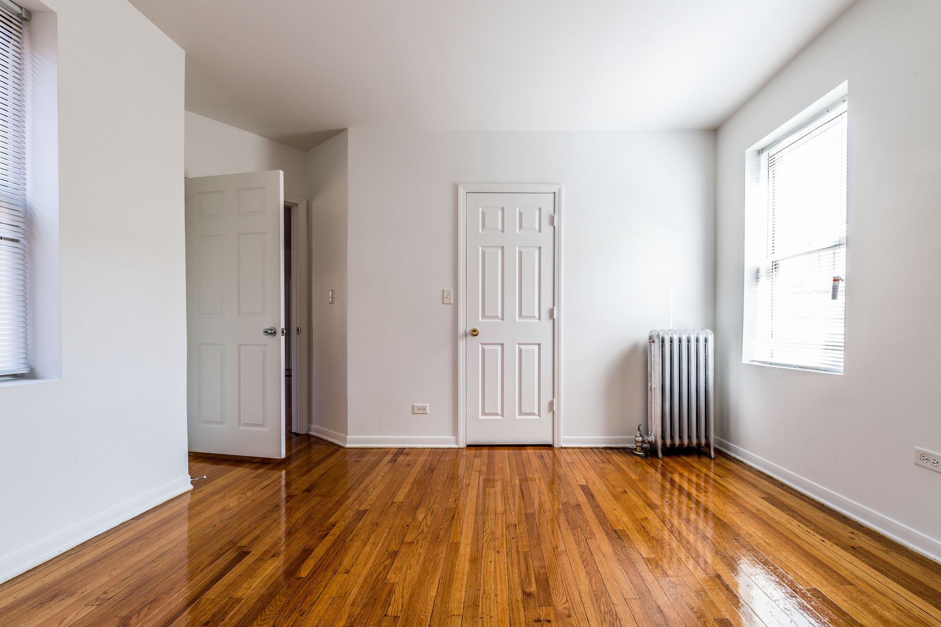Empty room with hardwood floors, white walls, two windows, and two white doors.