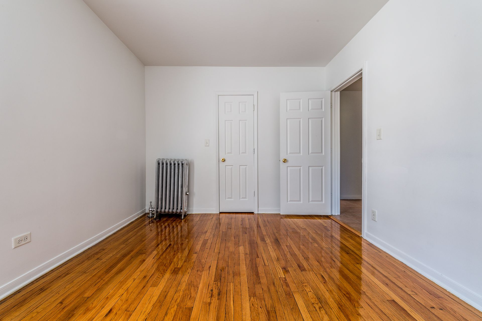 Empty room with hardwood floors, white walls, two doors, and a radiator.