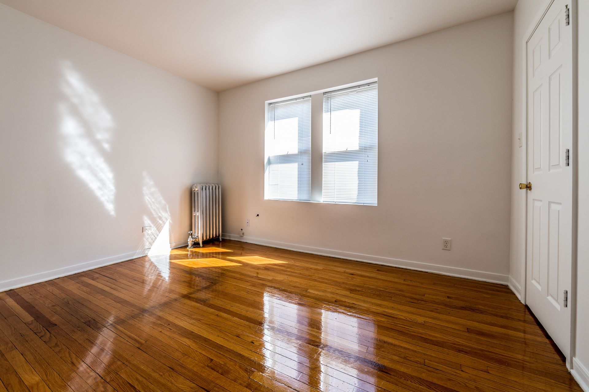 Empty room with hardwood floors, a window with blinds, and a radiator. Bright sunlight.