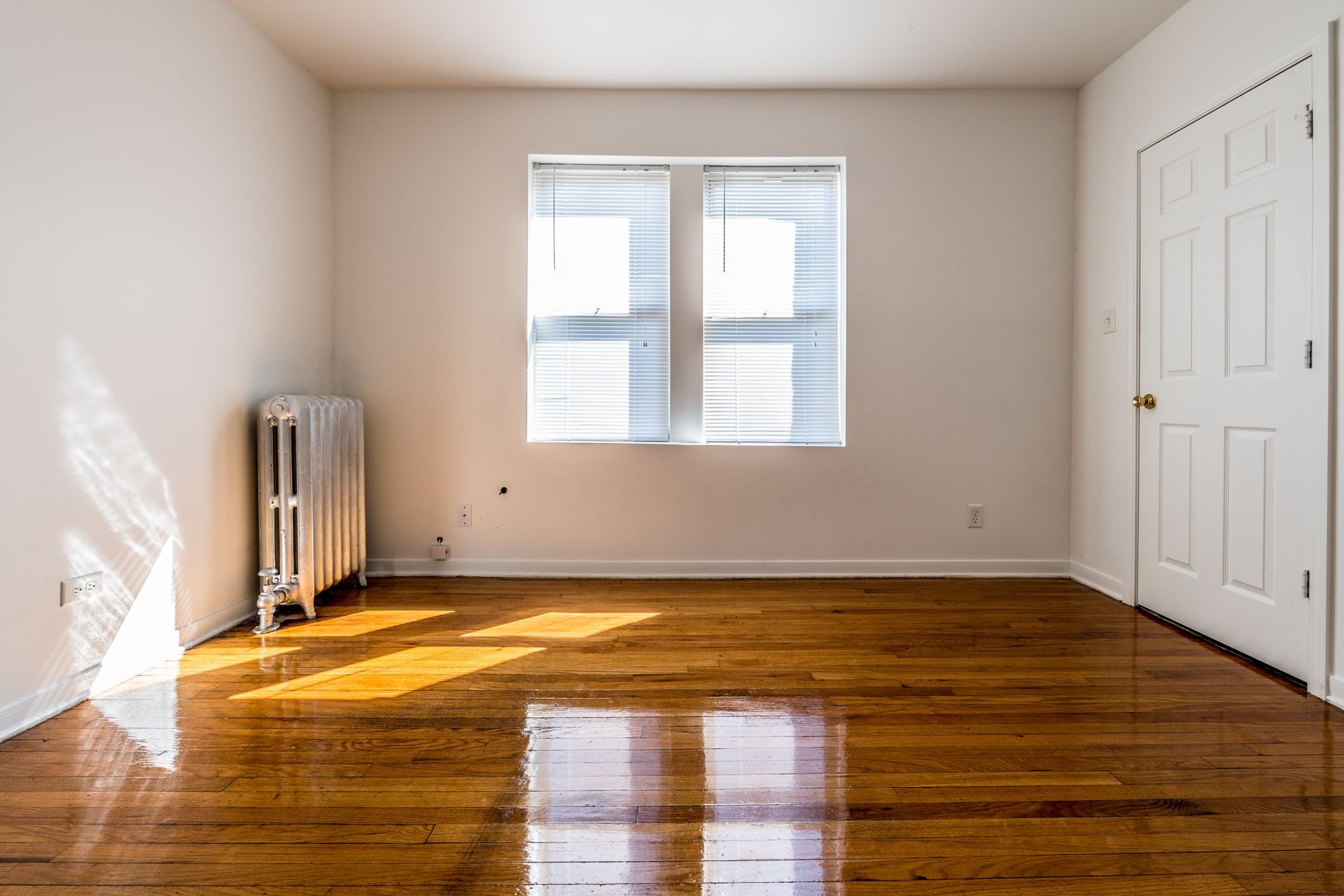 Empty room with hardwood floors, a window, and a radiator. Bright sunlight.