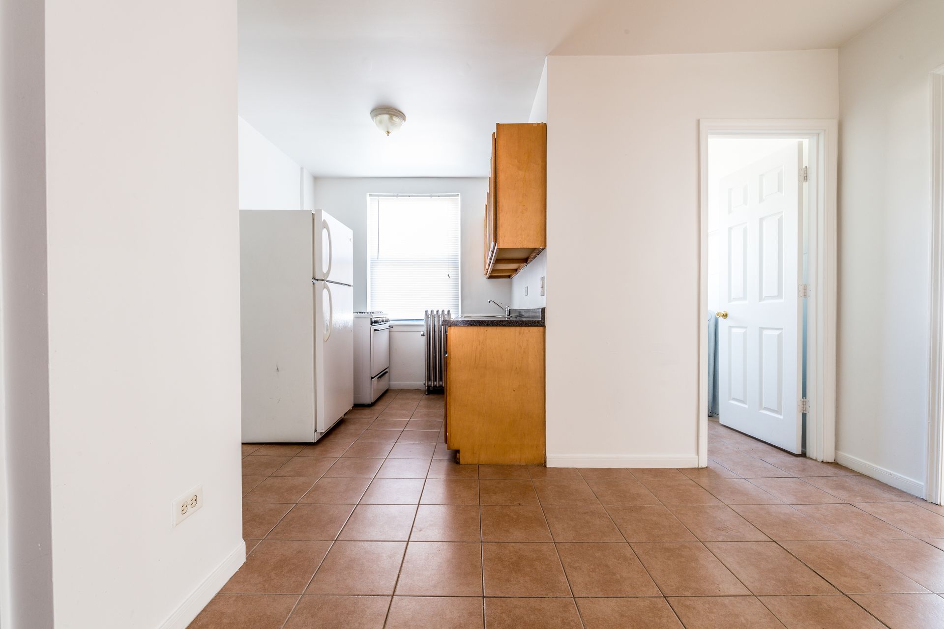 Empty kitchen with white appliances, wooden cabinets, and tan tile floor. Open doorway to a white door.