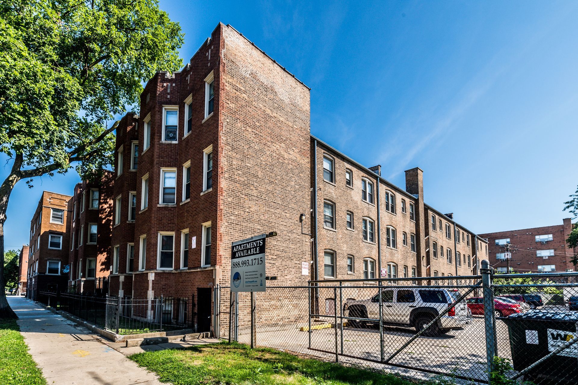 Brick apartment building with parked cars behind a chain-link fence on a sunny day.