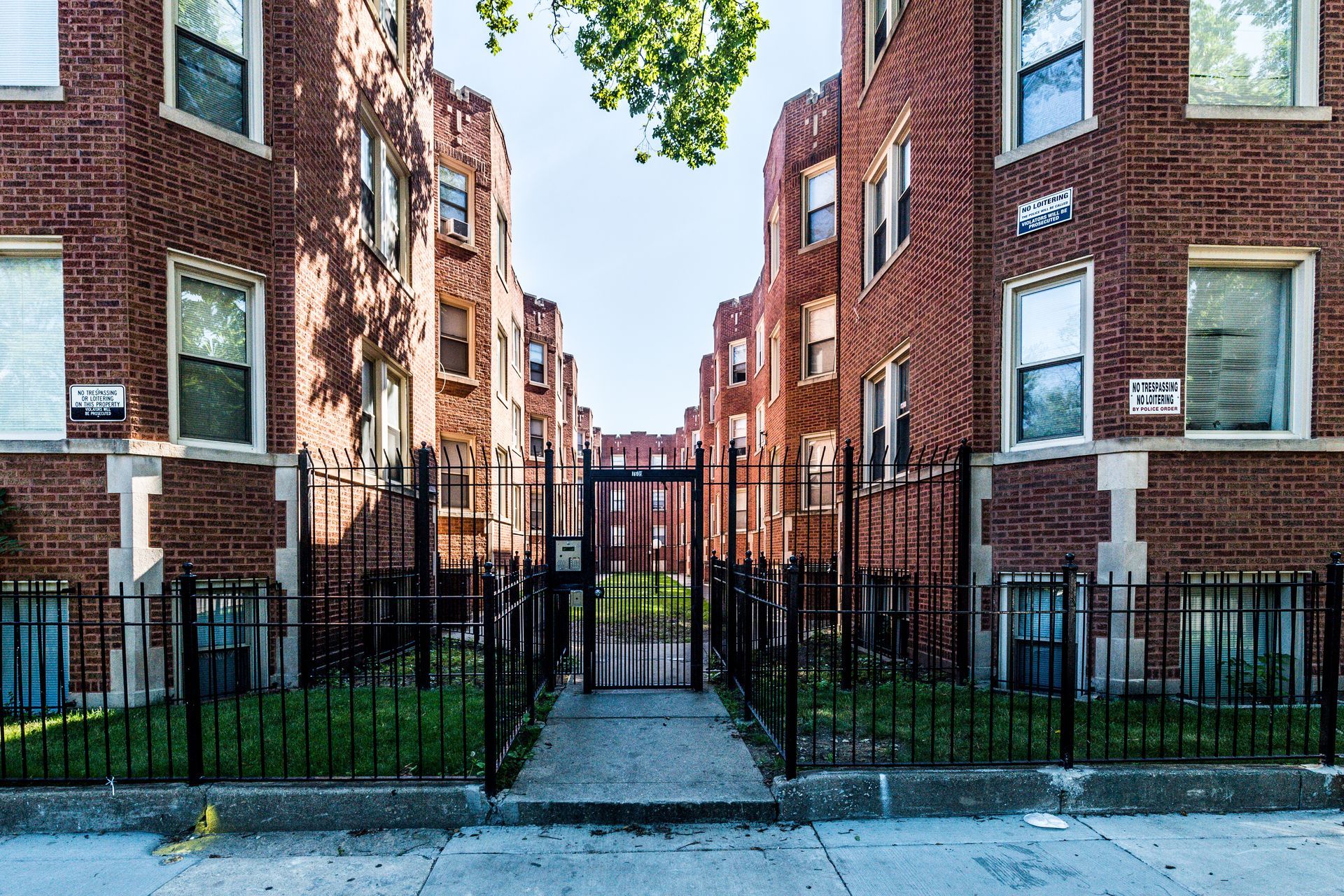 Brick apartment buildings with a black wrought iron fence and gate, a grassy courtyard visible beyond.