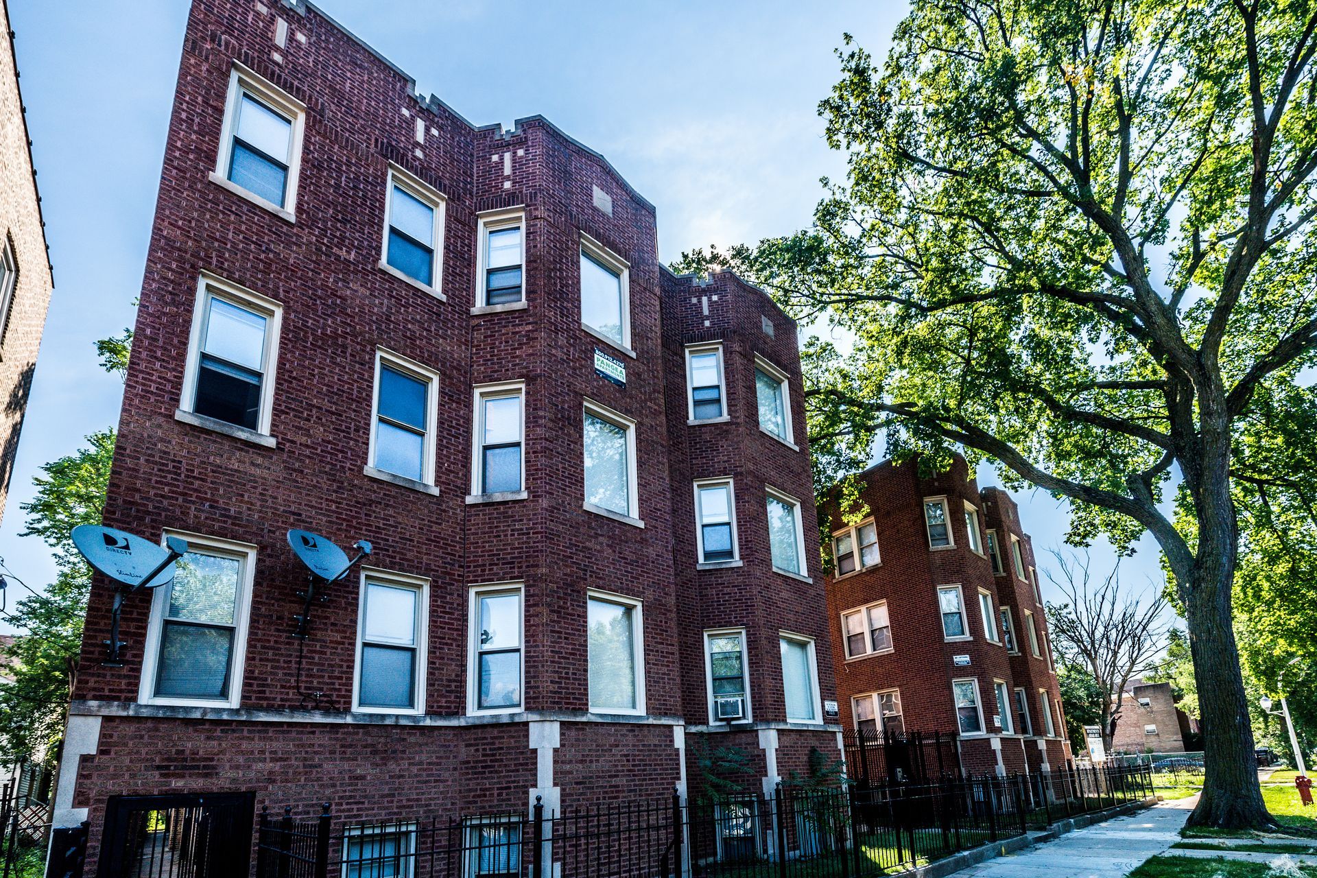 Red brick apartment buildings with satellite dishes, next to a large tree and sidewalk.