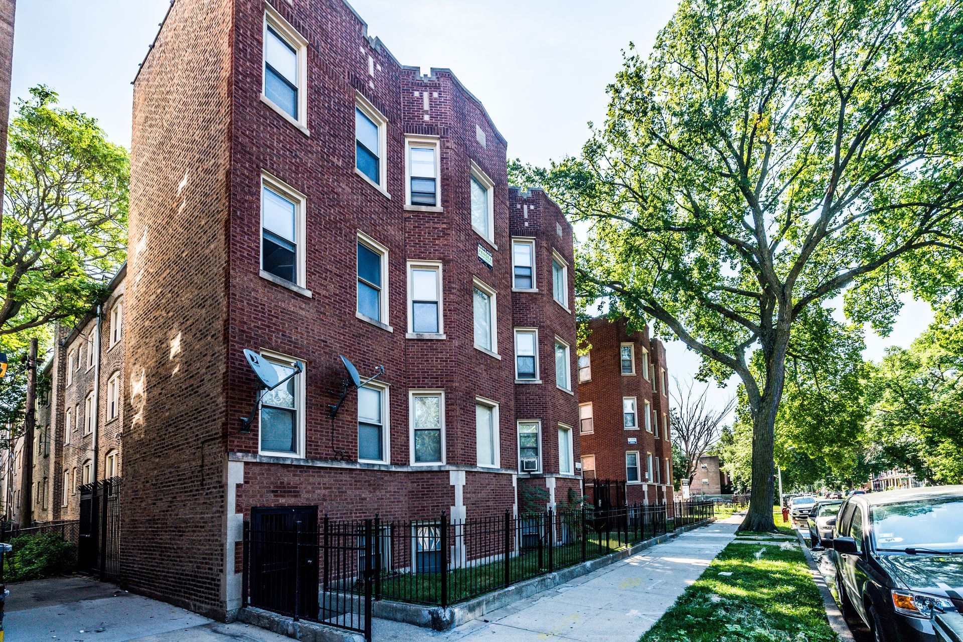 Brick apartment building on a tree-lined street with cars parked along the curb.