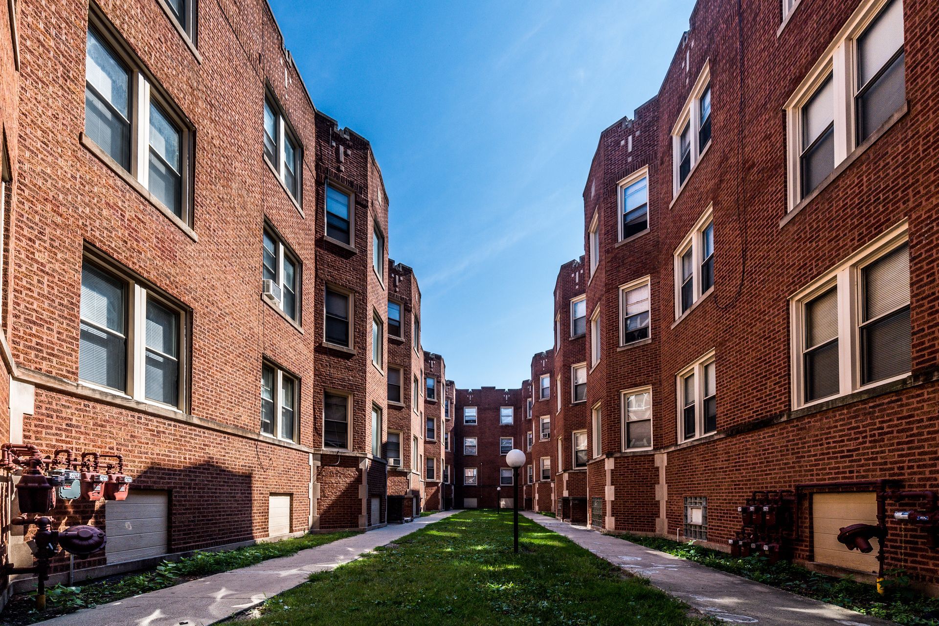 Brick apartment buildings face each other, forming a grassy courtyard under a blue sky.