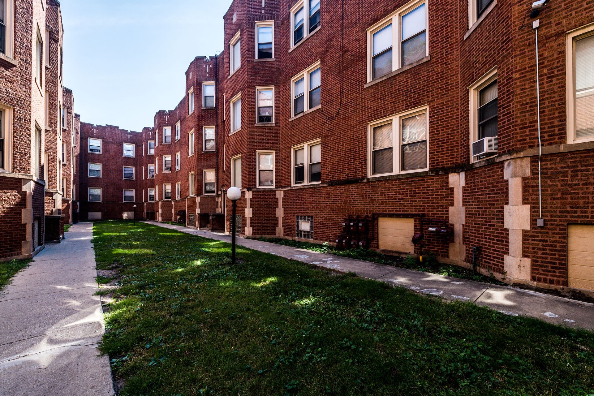 Apartment buildings with red brick exterior, windows, and a grassy courtyard with a sidewalk and lamp.