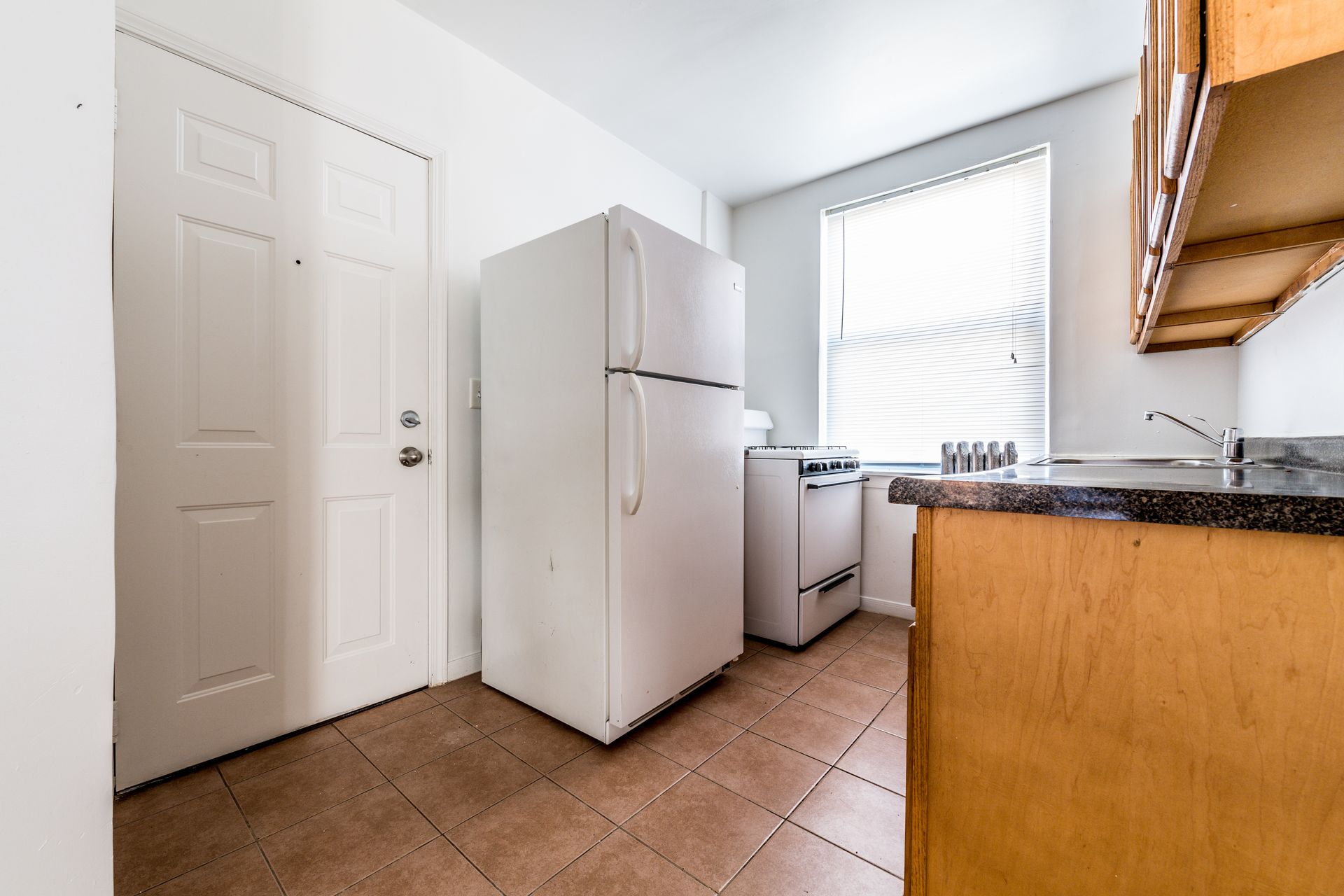 Small kitchen with a refrigerator, stove, cabinets, and a door. Light brown tile flooring.