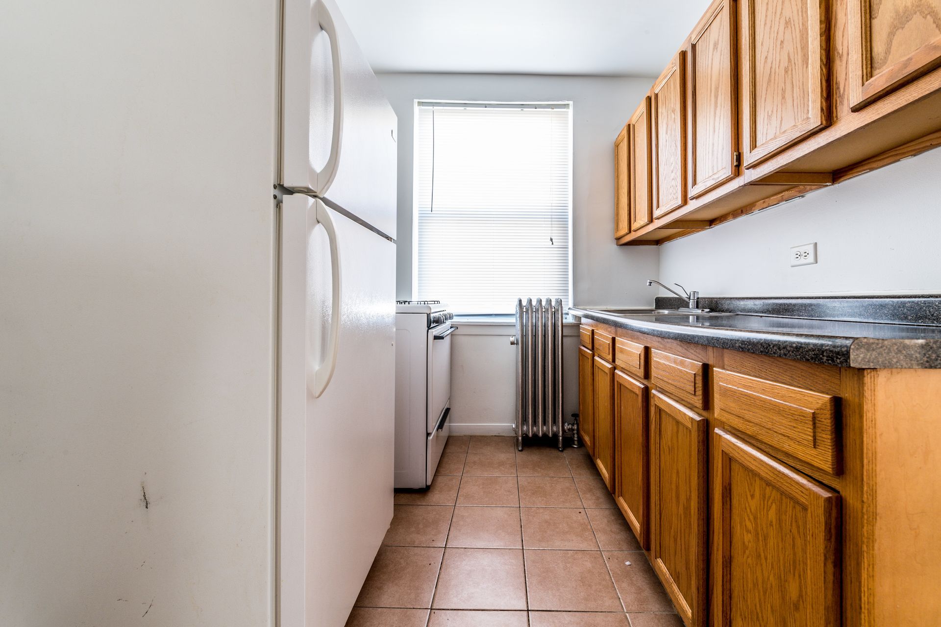 Small kitchen with white appliances, wooden cabinets, and a window with blinds.