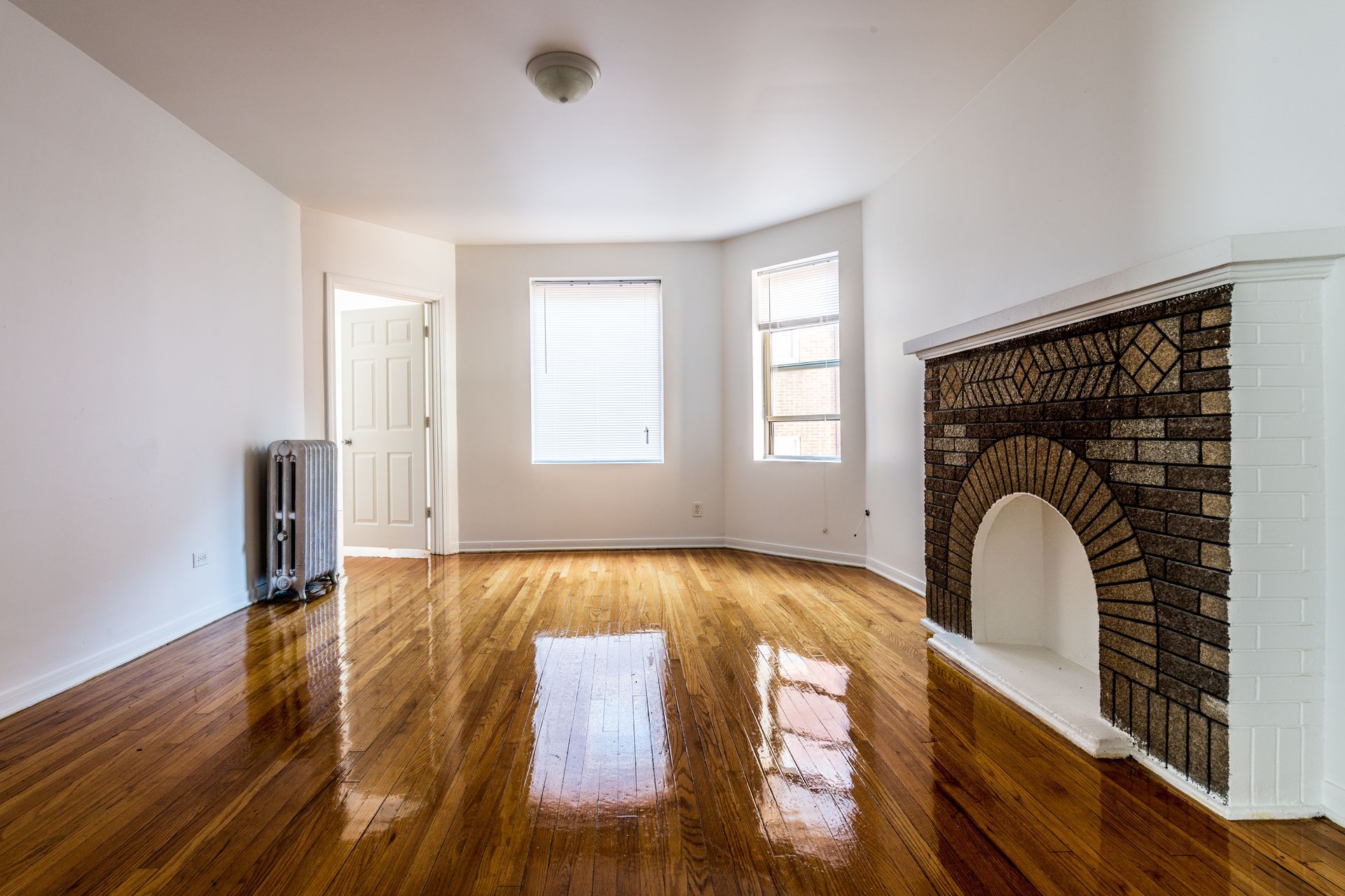 Empty room with hardwood floors, windows, and a brick fireplace.
