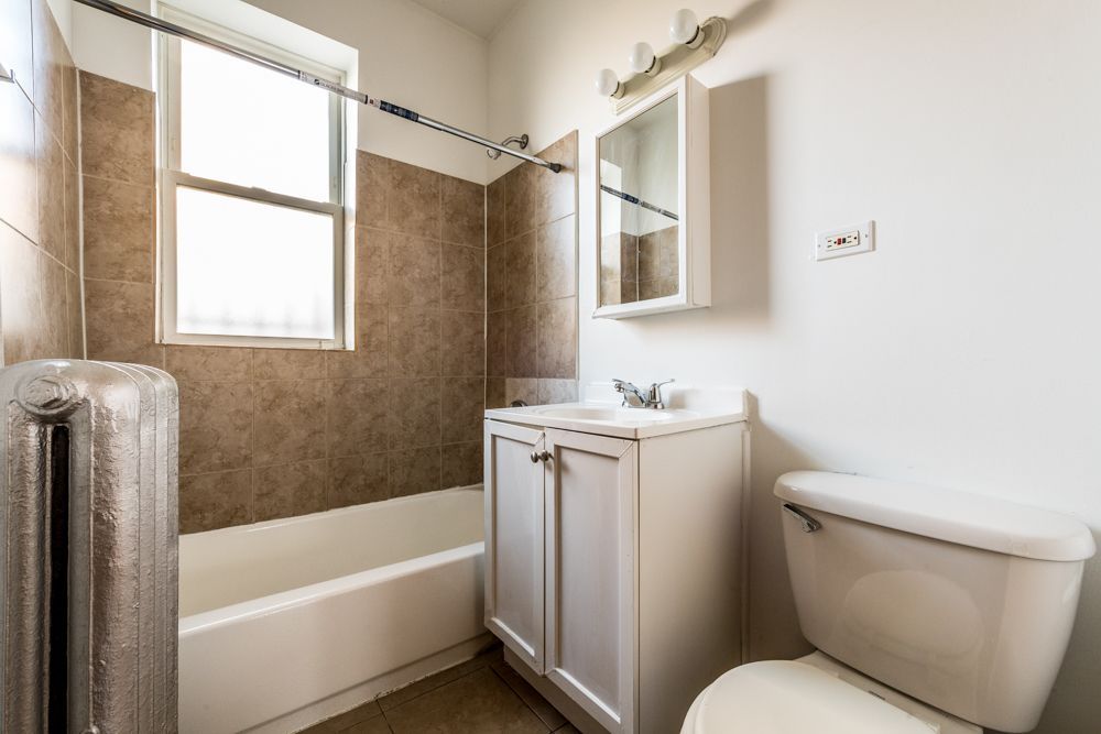 Bathroom with white fixtures, brown tile in shower, small window, and radiator.