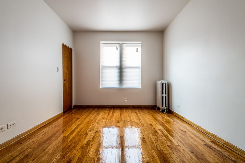 Empty room with wooden floors, white walls, window with blinds, radiator, and closed wooden door.