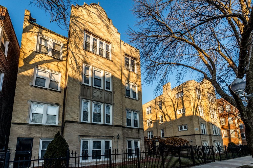 Three-story apartment building with tan brick exterior; bare tree in foreground, sunny day.