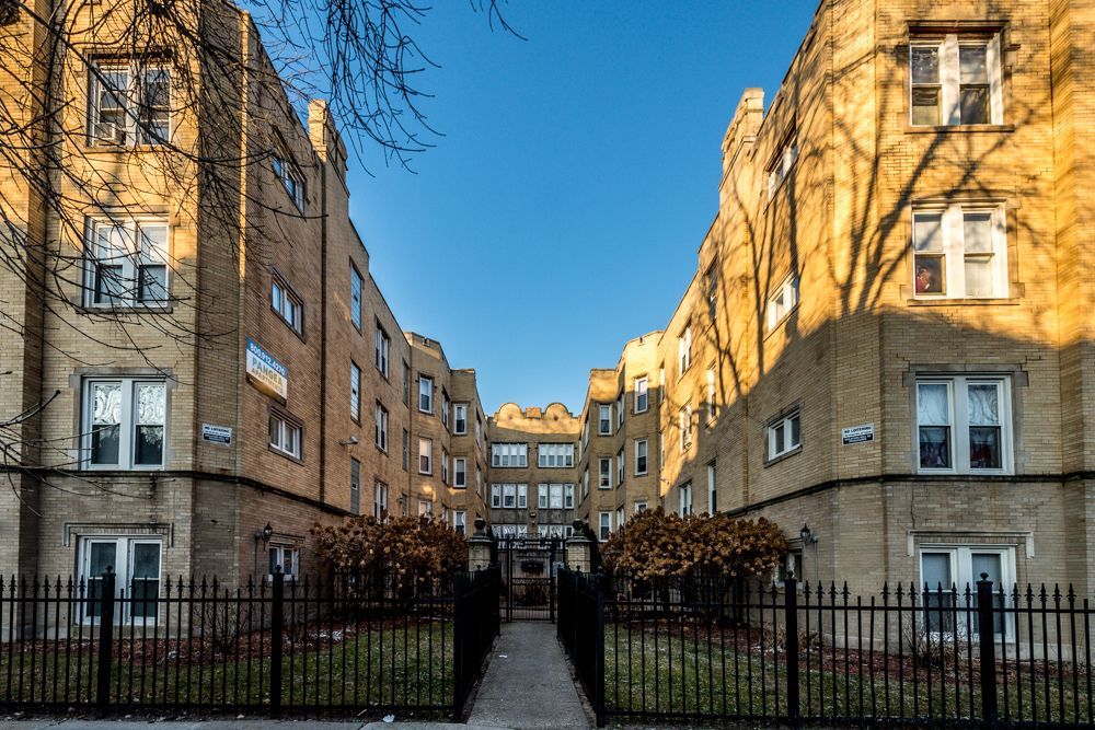 Apartment buildings with beige brick facades, black fence, walkway, and blue sky.