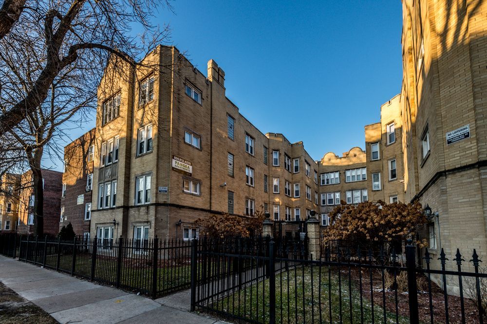Brown brick apartment building with wrought-iron fence, sidewalk, and trees under a clear blue sky.