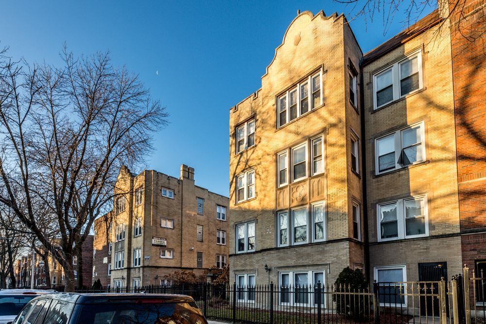 Apartment buildings with tan brick exteriors, clear sky. Cars parked on street, bare trees in foreground.