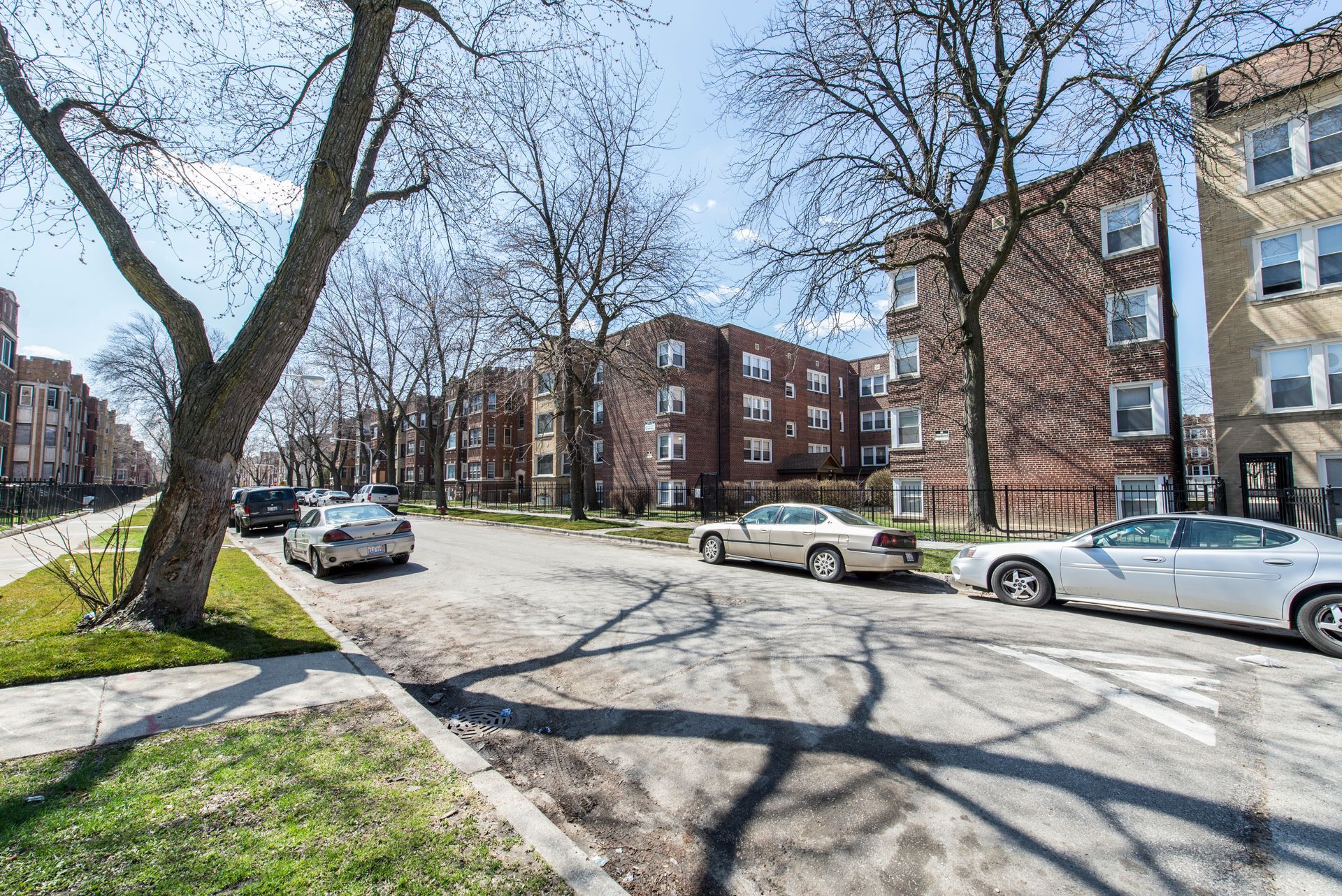 Street view with parked cars, apartment buildings, and bare trees under a bright sky.