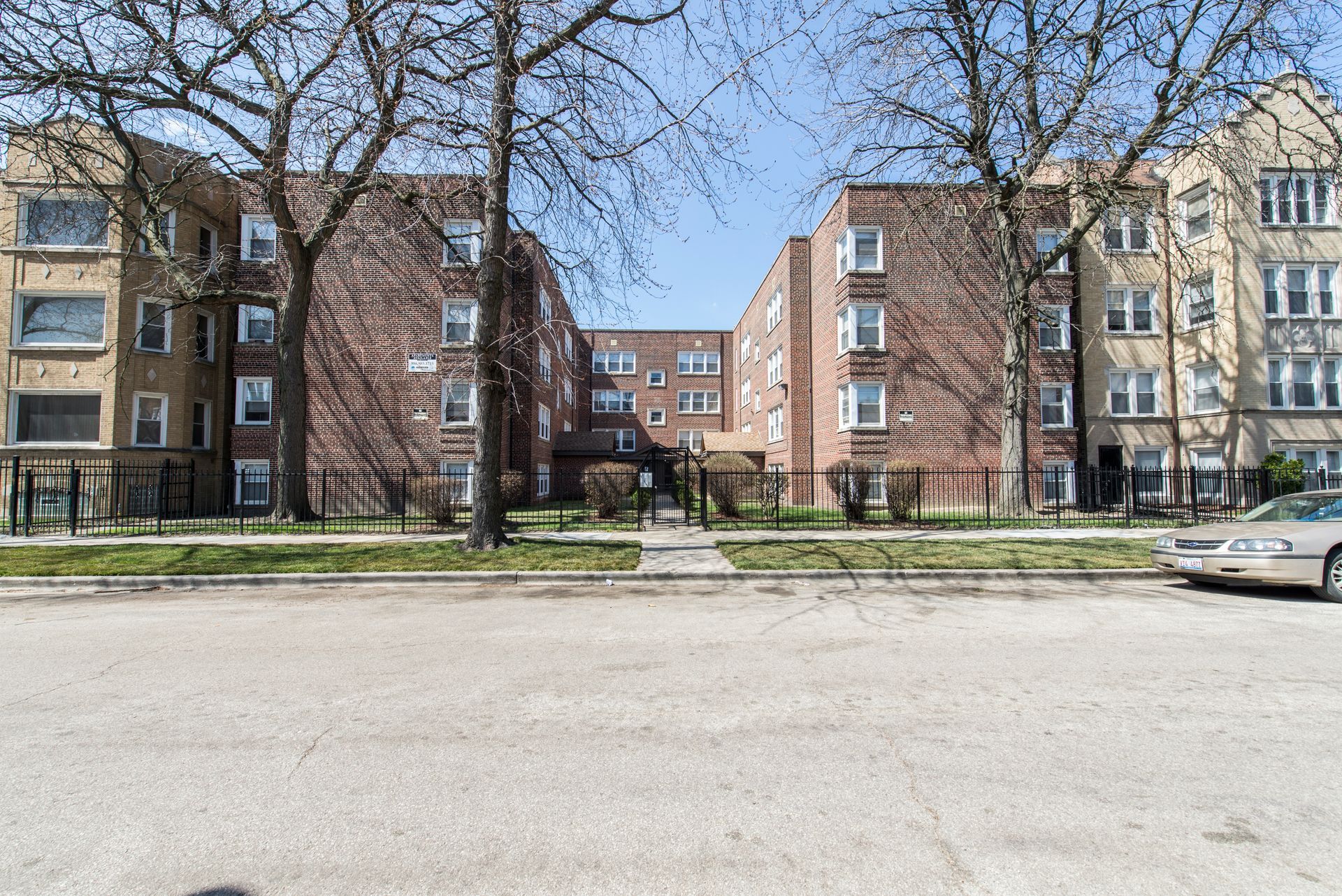 Brick apartment buildings with trees in front, viewed from a street on a sunny day.