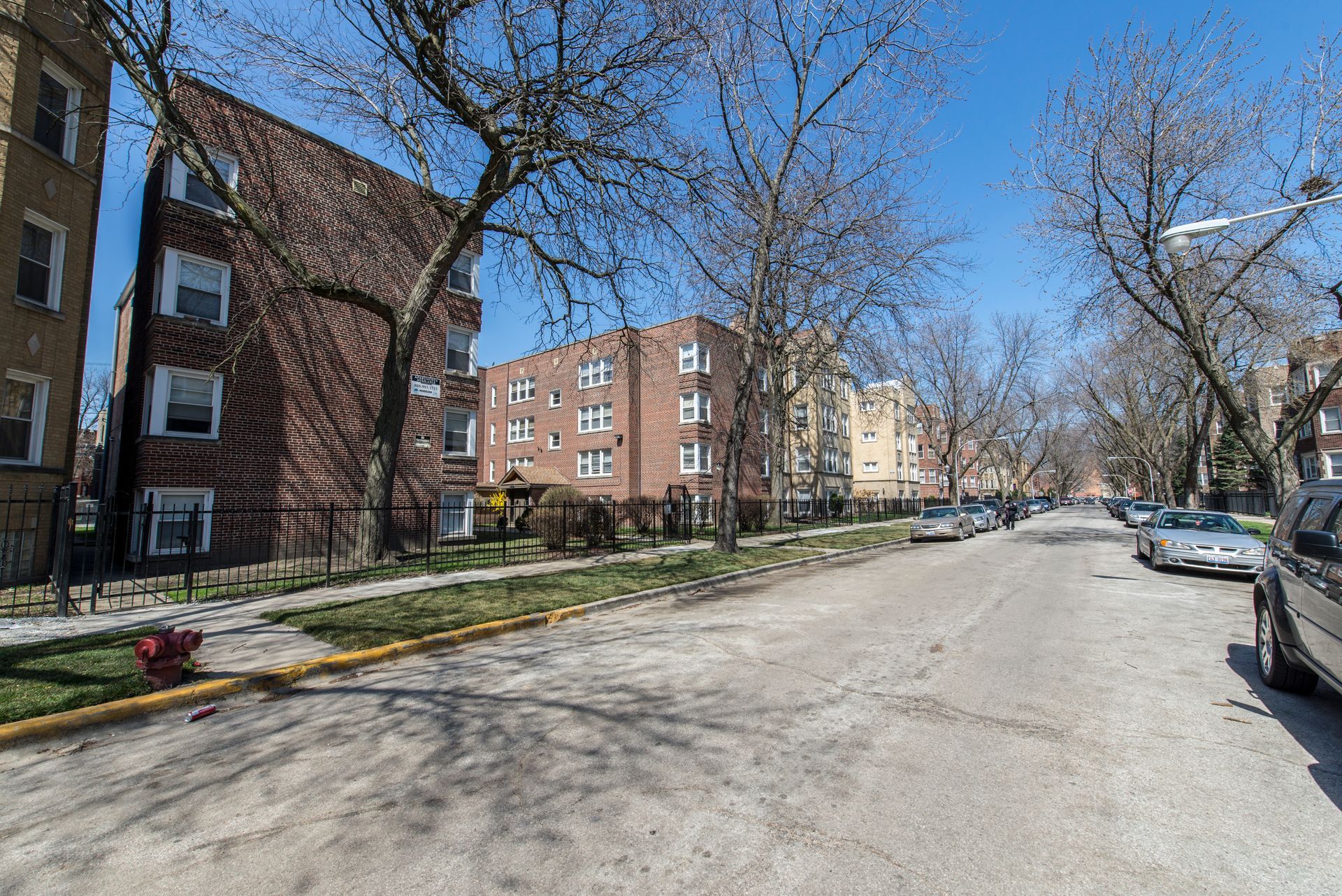 Street view of apartment buildings on a sunny day with parked cars. Bare trees line the sidewalk.