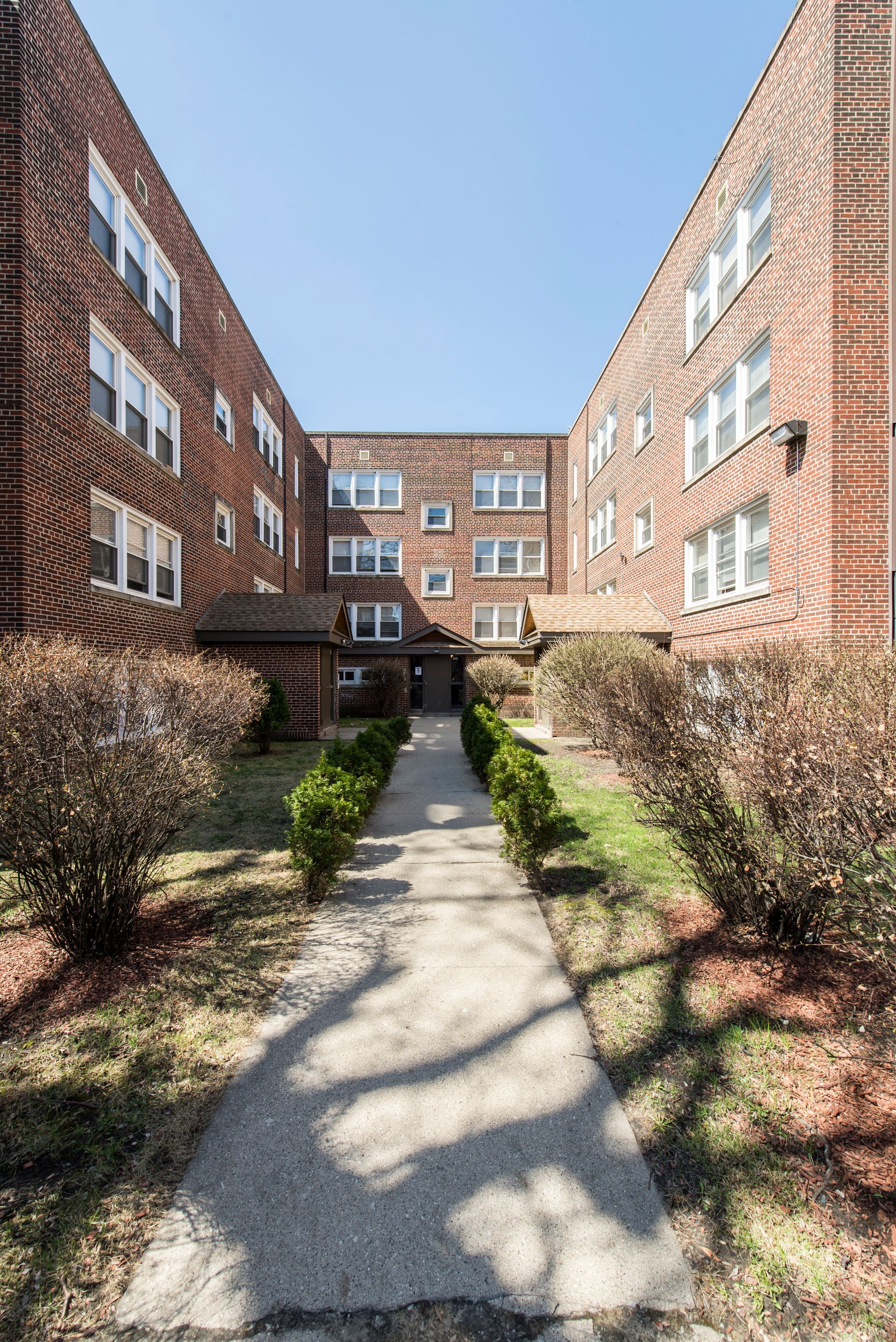 Red brick apartment building with a pathway leading to the entrance. Shrubs line the walkway.